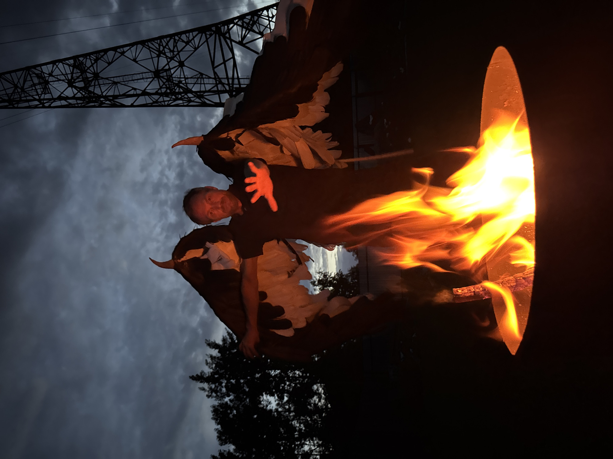 A medium shot shows a man in a dark t-shirt standing in front of a fire pit with large, feathered wings attached to his back. The man's right hand is outstretched, glowing red from the fire. The wings are dark brown on the upper portion, with white feathers towards the bottom. A tall, metal electrical tower looms in the background, silhouetted against a dark, cloudy sky. Trees and a fence are visible behind the man. The scene appears to be outdoors at dusk. This image is not identifiable as being in Chaikovsky, Russia.