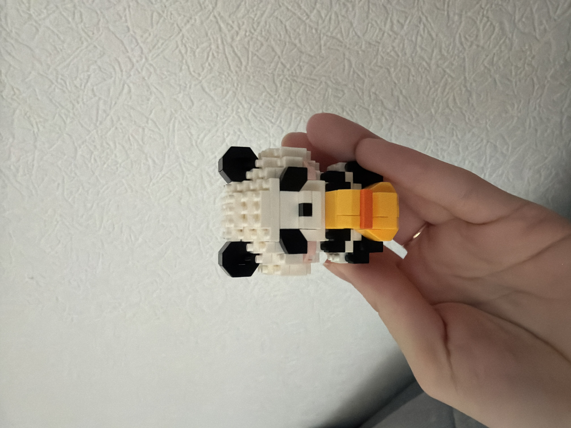 A hand holds a miniature panda figurine made of building blocks. The panda is white with black accents on its ears, eyes, and limbs, and it wears a yellow and orange outfit. The background is a textured white wall. The image was taken in Kyiv, Ukraine.