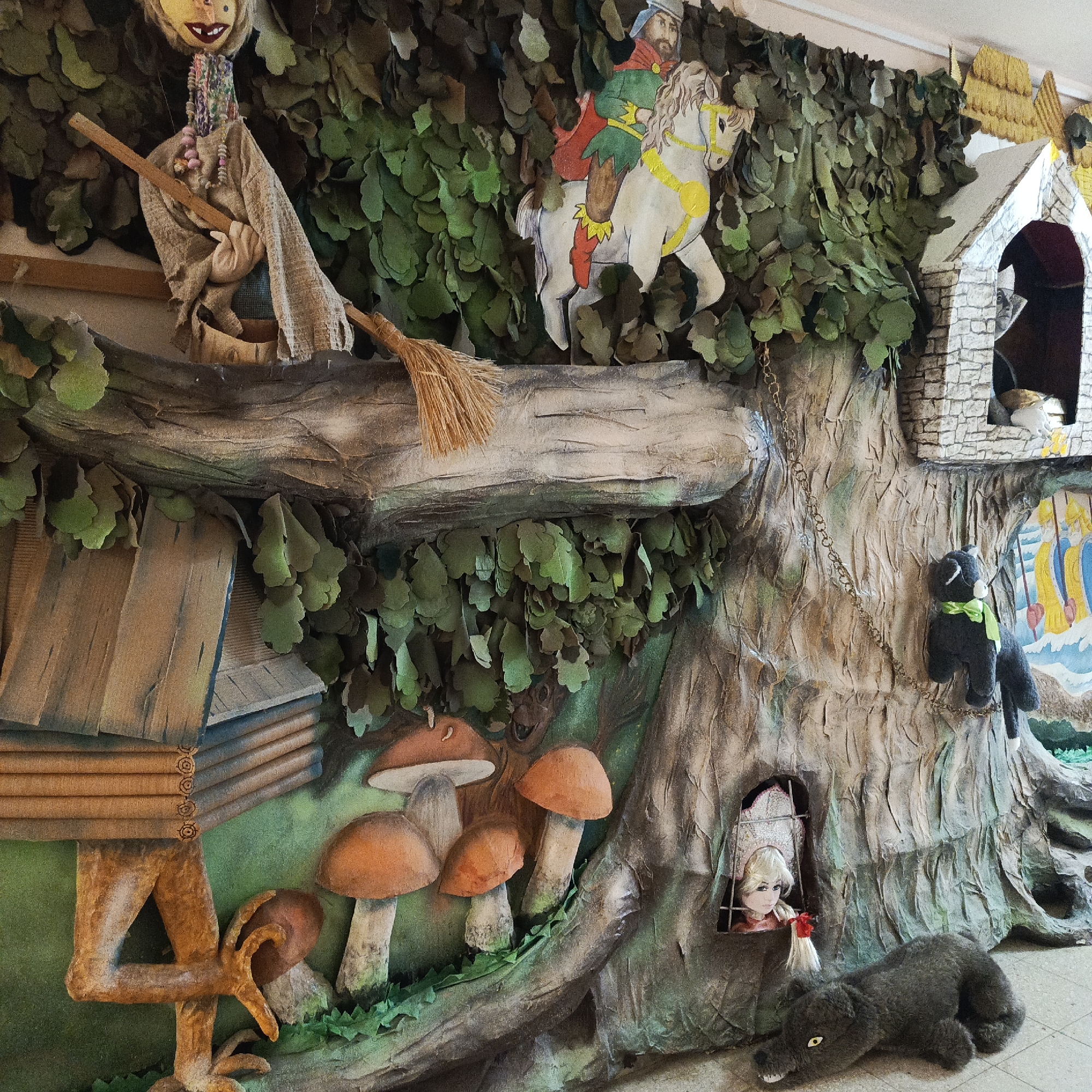The image shows a scene in Markovskii, Russia, possibly a theatrical stage set or children's exhibit, displaying characters and objects associated with Russian folklore. From left to right, there is a Baba Yaga figure in a mortar with a broom, a wooden house on chicken legs, and various mushrooms. Further right is a drawing of a knight on horseback, and a tree with a small stone building at the top. A doll is in a lower tree window, and a stuffed wolf is lying in front of the tree. Hanging from the tree is a stuffed cat with a green scarf.