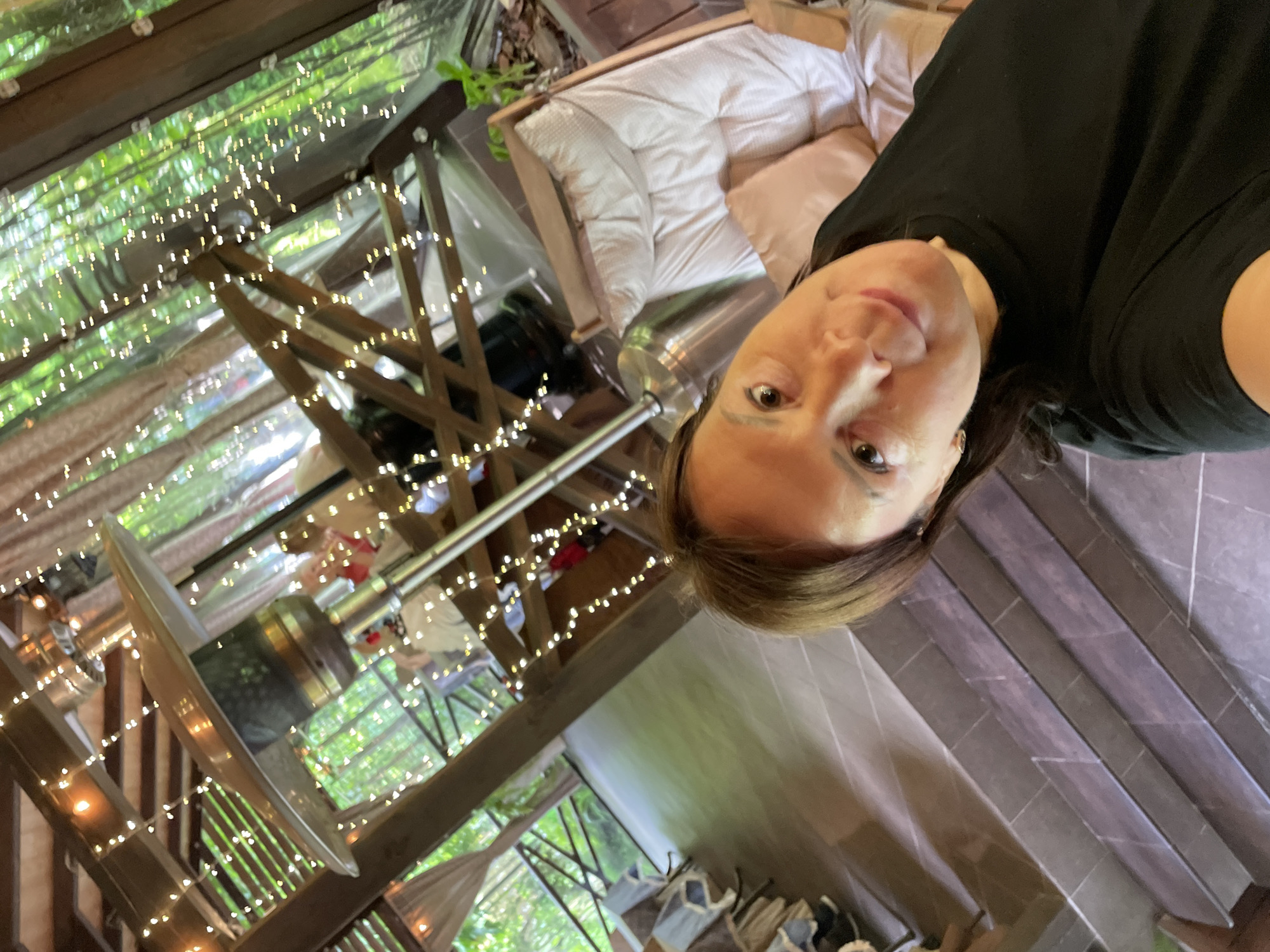 A woman wearing a black t-shirt is taking a selfie inside a gazebo in Solokh-Aul, Russia. The gazebo has a wooden frame decorated with string lights. Inside, there is a daybed with white bedding and a patio heater. The exterior of the gazebo is visible through the sides, revealing greenery and a hillside. The flooring near the woman appears to be gray tiles.