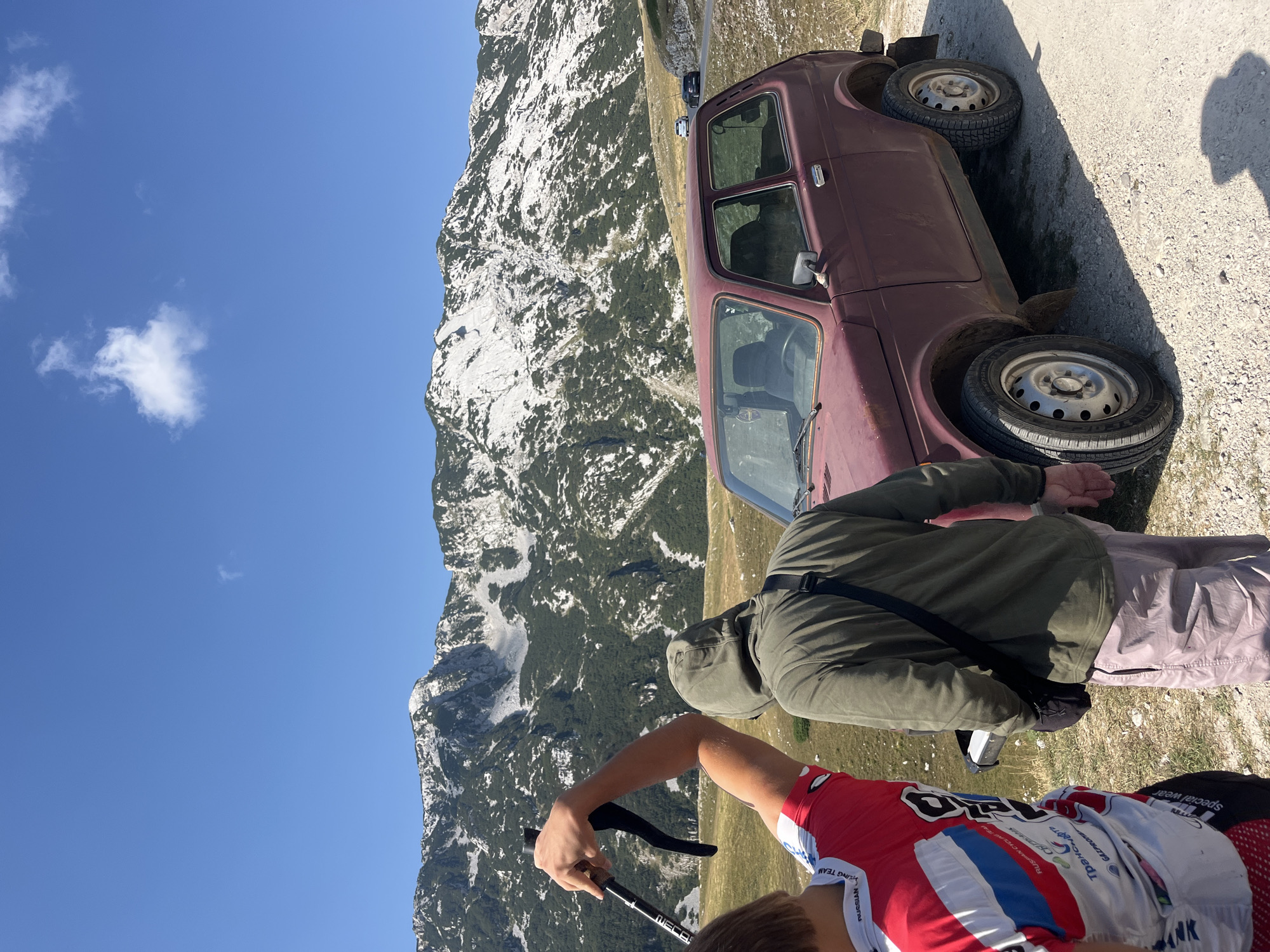 A burgundy Lada Niva is parked on a gravel road in a mountainous area. Two individuals are standing near the car, facing away from the camera. The person on the left is wearing a red, white, and blue cycling jersey and holding a hiking pole. The person on the right is wearing a green hooded sweatshirt and light-colored pants. In the background, a range of mountains covered in sparse green vegetation and patches of snow stretches across the horizon under a clear blue sky with a few wispy clouds. The location is identified as being in the vicinity of Dubrovsko, Montenegro.