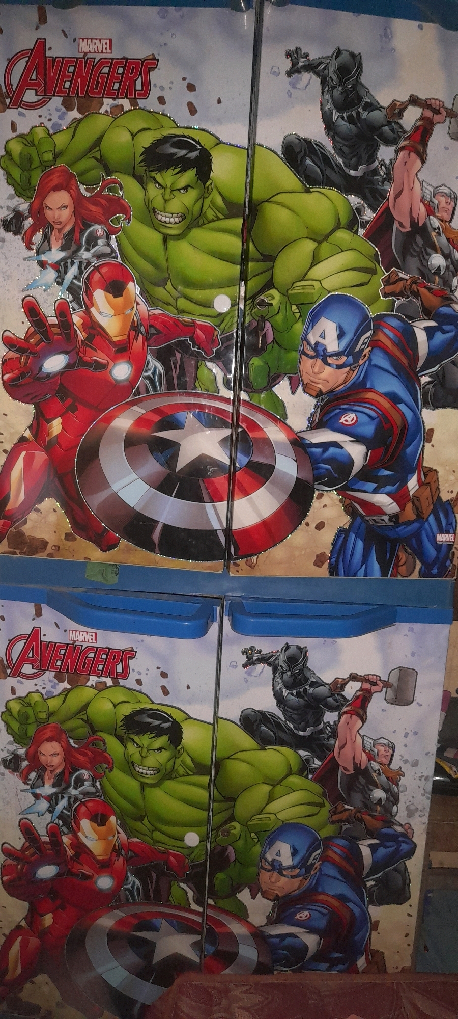 The image shows a close-up, eye-level shot of two identical, stacked, blue plastic storage bins with hinged lids, featuring a Marvel Avengers comic-style graphic design. The bins are placed side-by-side. The graphic depicts a dynamic group of Avengers characters in action poses against a speckled background.
On the top bin, from left to right, are: Black Widow, her body angled to the left, holding a weapon. Next to her is a large, muscular green Hulk, mid-action with a clenched fist. To the right of the Hulk, partially obscured, is Black Panther in a crouched, ready-to-pounce pose. To the right of Black Panther is Thor, holding his hammer aloft. In the foreground, on the left, is Iron Man in his red and gold suit, extending his left hand forward with his palm open, glowing. In front of Iron Man and partially overlapping is Captain America, holding his iconic shield, his body turned towards the right. His shield is prominently displayed.
The bottom bin repeats the same Avengers graphic, with the characters appearing slightly lower and wider. Again, Black Widow is on the left, followed by the Hulk, then Black Panther wielding his hammer. Iron Man is on the left in the foreground, and Captain America is on the right, also holding his shield. The graphics on both bins have a glossy, slightly textured finish, with some subtle glitter effects.
The environment appears to be indoors, with a hint of a cluttered background visible behind the bins. The bins themselves are the primary focus, filling most of the frame. There is no indication of the city of Tegal, Indonesia, within the image itself.