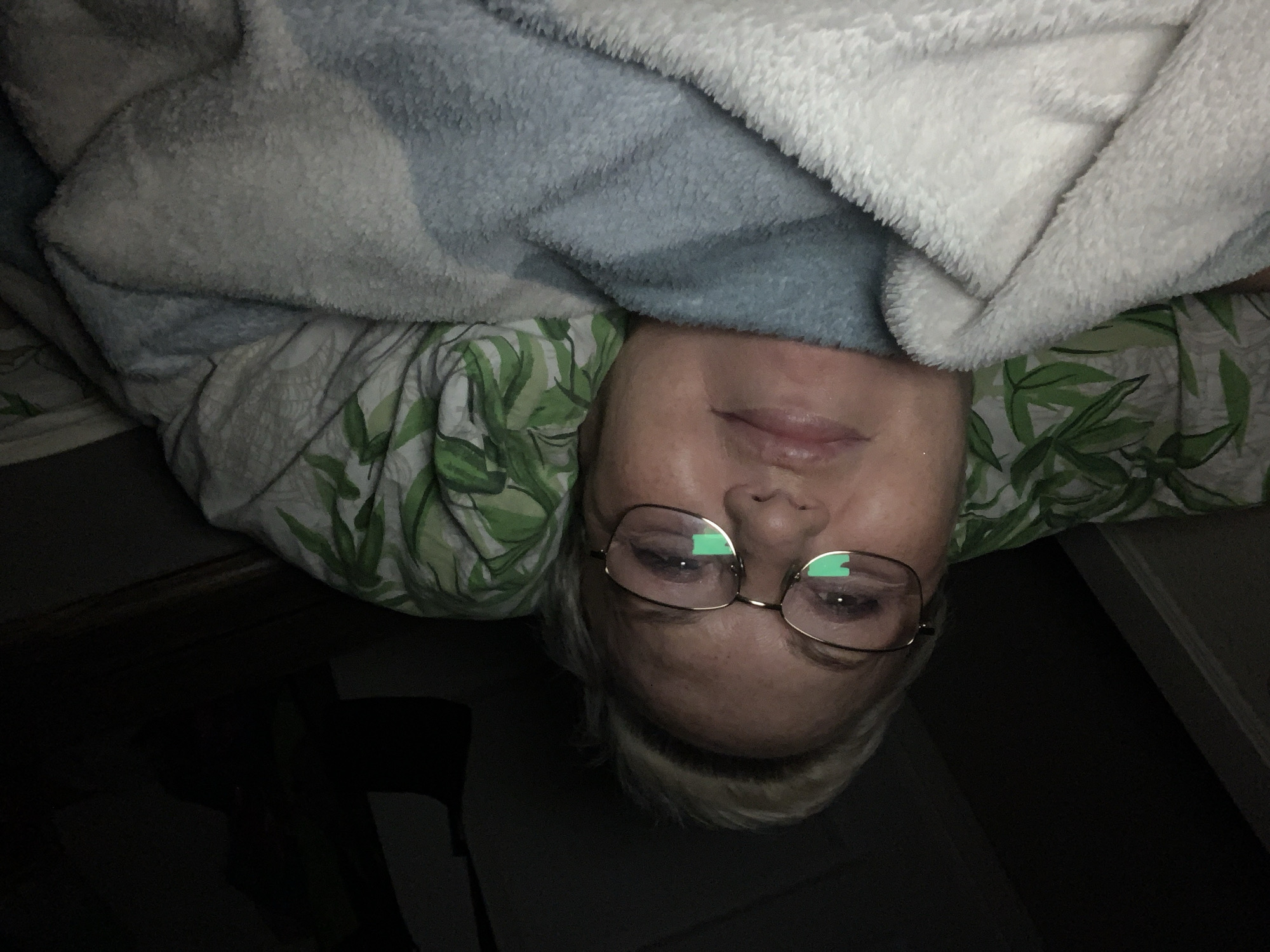An upside-down, indoor image shows a person wearing glasses under a blanket. The blanket appears to be made of white and blue fleece. The person is also beneath a green and white patterned bedsheet. A dark area is in the lower portion of the image. It is not possible to determine the location based on the image provided.