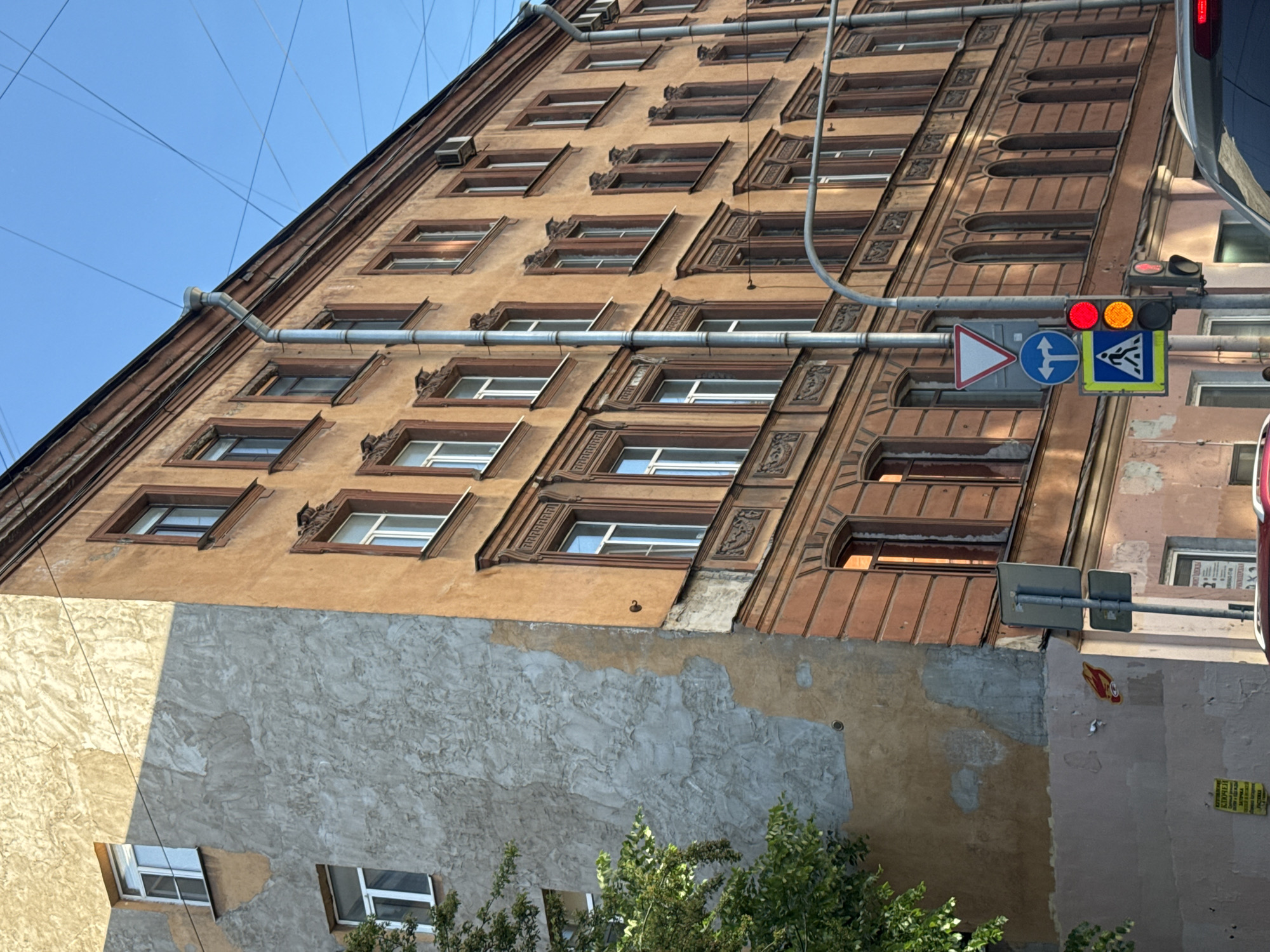 This is a medium shot, taken outdoors, of an old, multi-story building in Saint Petersburg, Russia. The building has a tan-orange facade with decorative trim around the windows. On the left side of the building, part of the facade is a rough, light-gray plaster that is peeling away. There are several windows with white frames on both sections of the building, and some have a faint orange glow from inside. A drainpipe runs vertically down the tan-orange facade. In the background, a clear blue sky is visible with a web of overhead electrical wires. A street-level view shows traffic lights, street signs, and a small section of a car's roof. A tree with green foliage is visible on the far left of the image.