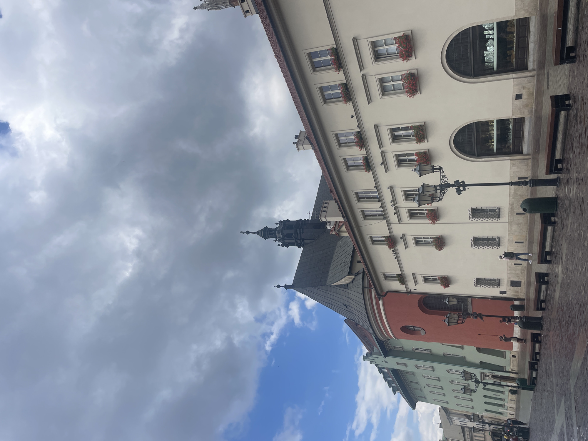The image depicts a cloudy day in Krakow, Poland, likely in the Old Town. A row of historic buildings lines a cobblestone square. The architecture features a variety of colors and styles, with a prominent red building with arched windows and a dark-steepled tower. The buildings are adorned with flower boxes containing red flowers in their windows. Several people are visible walking in the square, some carrying bags. Benches are placed along the square, and street lamps and trash receptacles are also present. The ground appears wet, suggesting recent rain.
