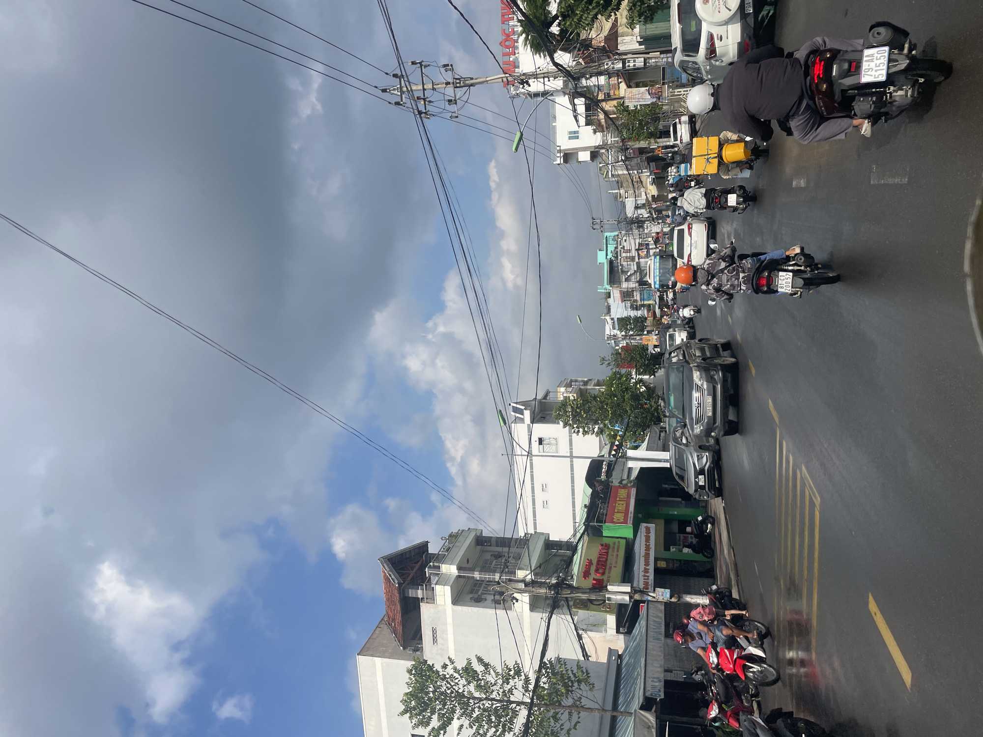 Captured from a slightly elevated perspective in Bac Nha Trang Ward, Vietnam, the image portrays a busy street scene. The street is wet, presumably from recent rain, and reflects the overcast sky with patches of blue breaking through. Overhead, a network of electrical wires crisscrosses the view.
On the right side of the street, a line of motorbikes and cars are visible. Several individuals are riding motorbikes. Shop signs, including "COM THIEN THANH" and "ĐẠI LÝ ỦY QUYỀN BẠC PHỦ QUY", are visible along the roadside. The buildings are a mix of residential and commercial structures. The license plate "81-C1 200.57" and "79-AA 515.50" are also visible on a motorbike. The image captures the everyday hustle and bustle of urban life in Nha Trang.