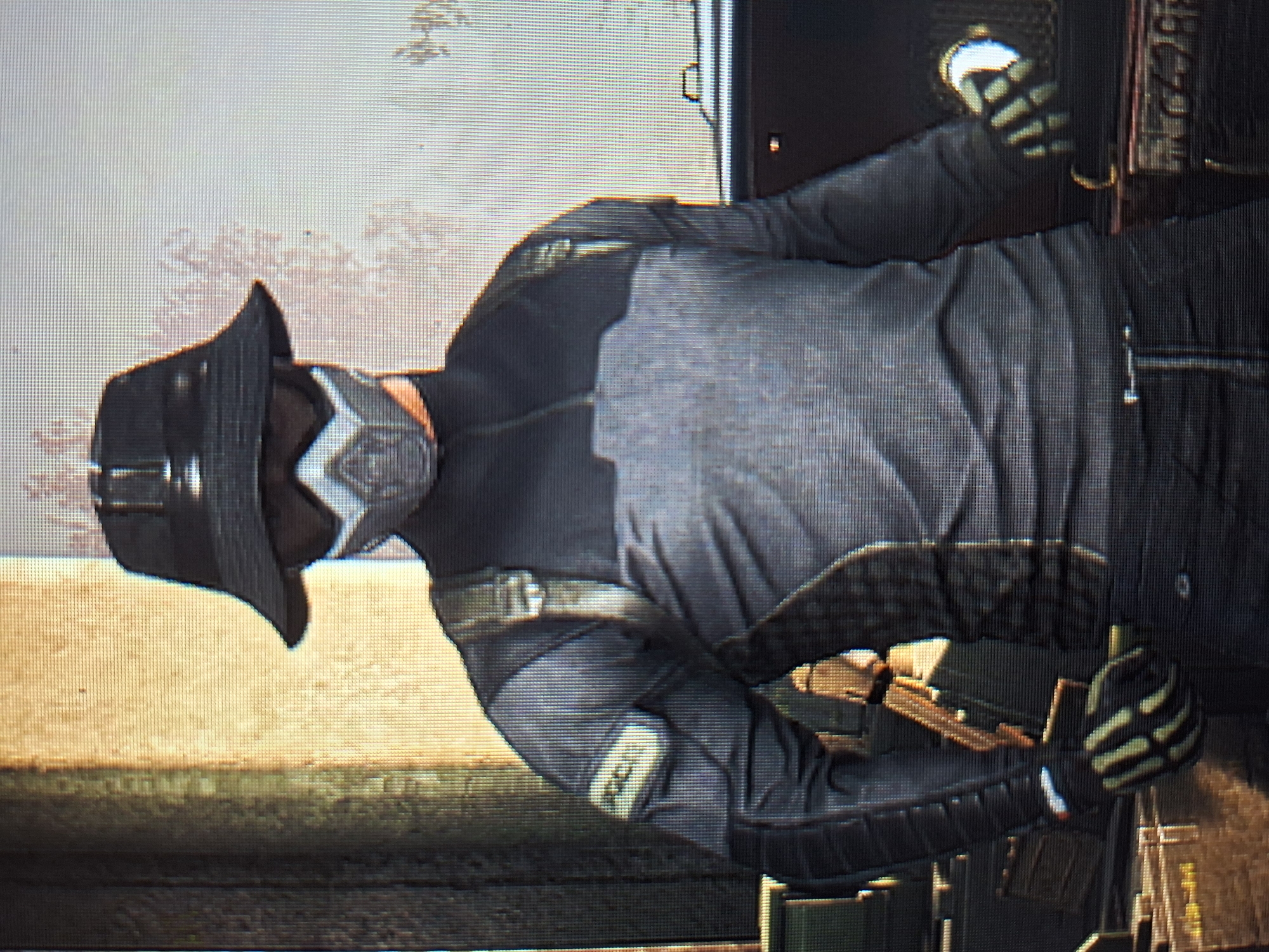 A first-person perspective shot shows a person wearing a dark bucket hat, a white and silver gas mask covering their mouth and nose, and a dark gray long-sleeved shirt with lighter gray panels across the chest and shoulders. The person also wears dark pants, dark gloves with light green finger designs, and a tan strap over their right shoulder. A patch with yellow lettering, partially obscured, is visible on the person's left upper arm. The background features a textured, tan-colored wall on the left and a light gray, abstract background on the right. The person appears to be standing or walking in an urban environment, possibly a street or alley, given the wall texture and the hints of objects in the background that could be part of a city scene.