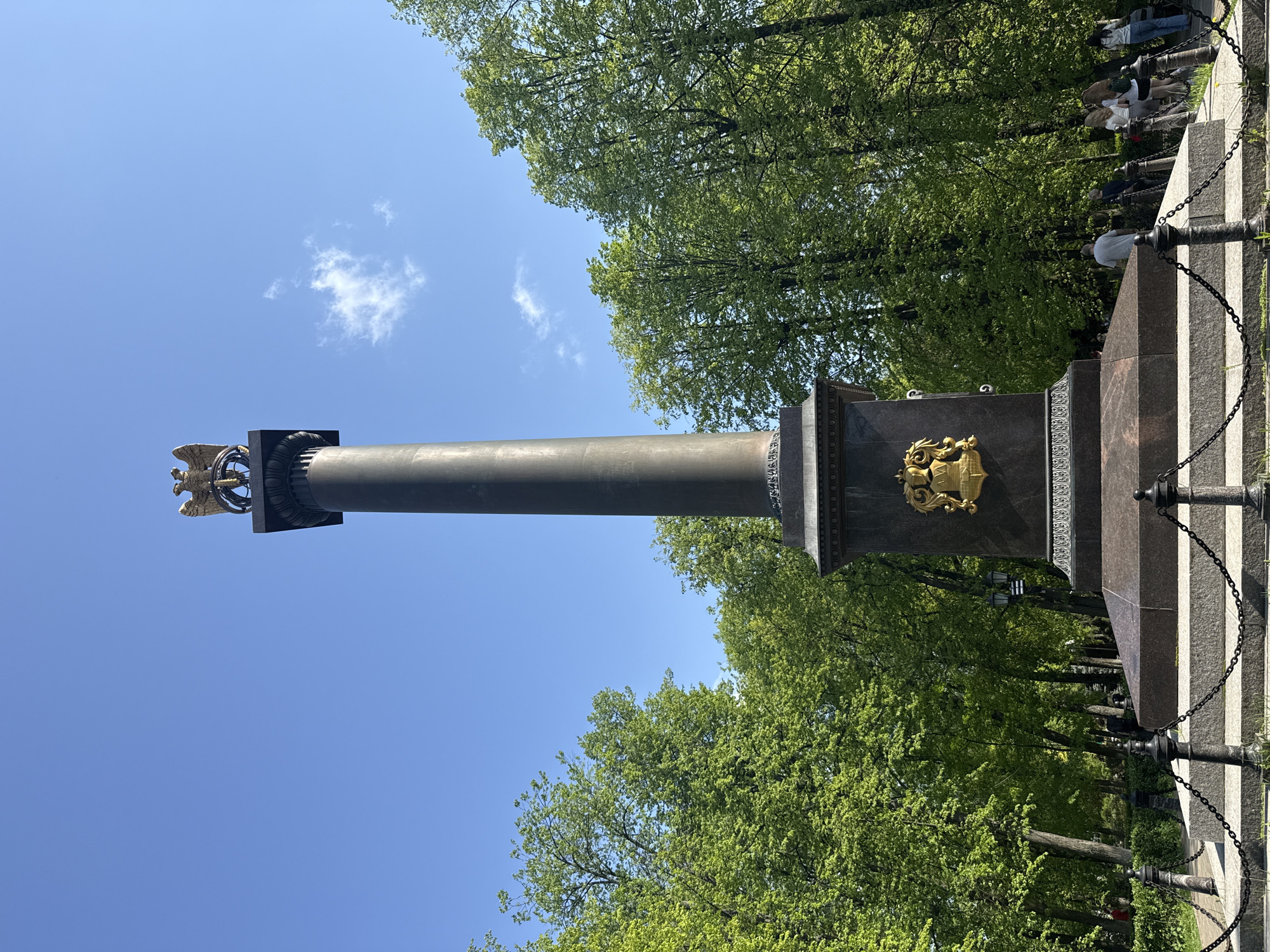 A low-angle, outdoor shot shows the Alexander Column in Заволжье, Russia. The column is tall and gray, topped with a golden eagle. It stands on a dark gray and red pedestal with a golden coat of arms on one side. Green trees surround the column, and a blue sky with some clouds is visible in the background. A black metal chain fence runs along the base. In the upper right corner, a few people are visible.