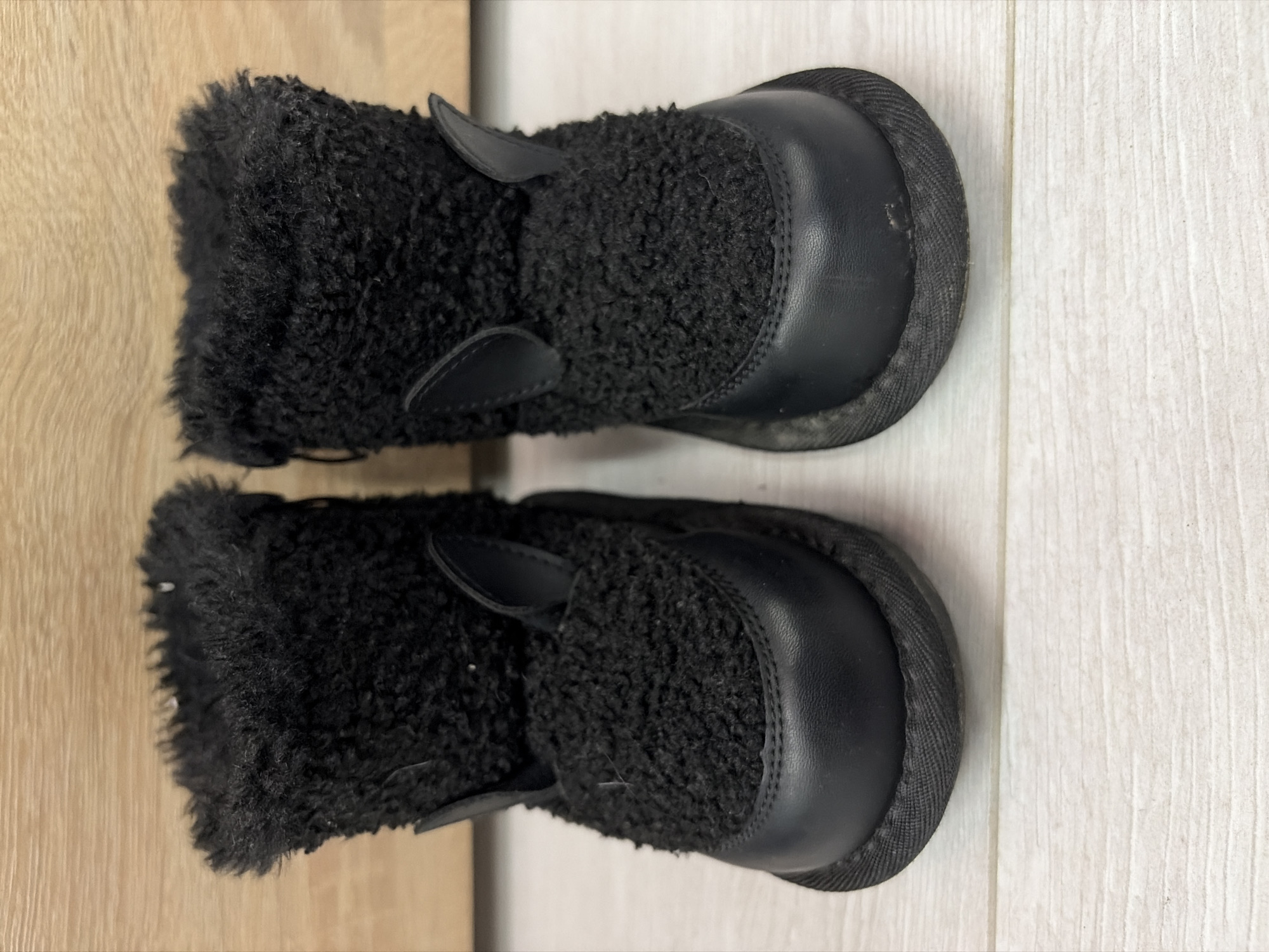 The image shows a pair of black, fur-lined boots on a wooden floor. The floor appears to be made of light-colored wooden planks on the right side and darker wooden planks on the left side. The boots are oriented vertically in the frame, with the toes pointing towards the top. The boots have a rounded toe cap made of a smooth, possibly leather-like material, while the rest of the boot is covered in a dense, black, curly fur. There are small, stylized "ears" or flaps protruding from the fur near the toe. The scene is indoors. There is no information on the city of Chaikovsky, Russia in the image.