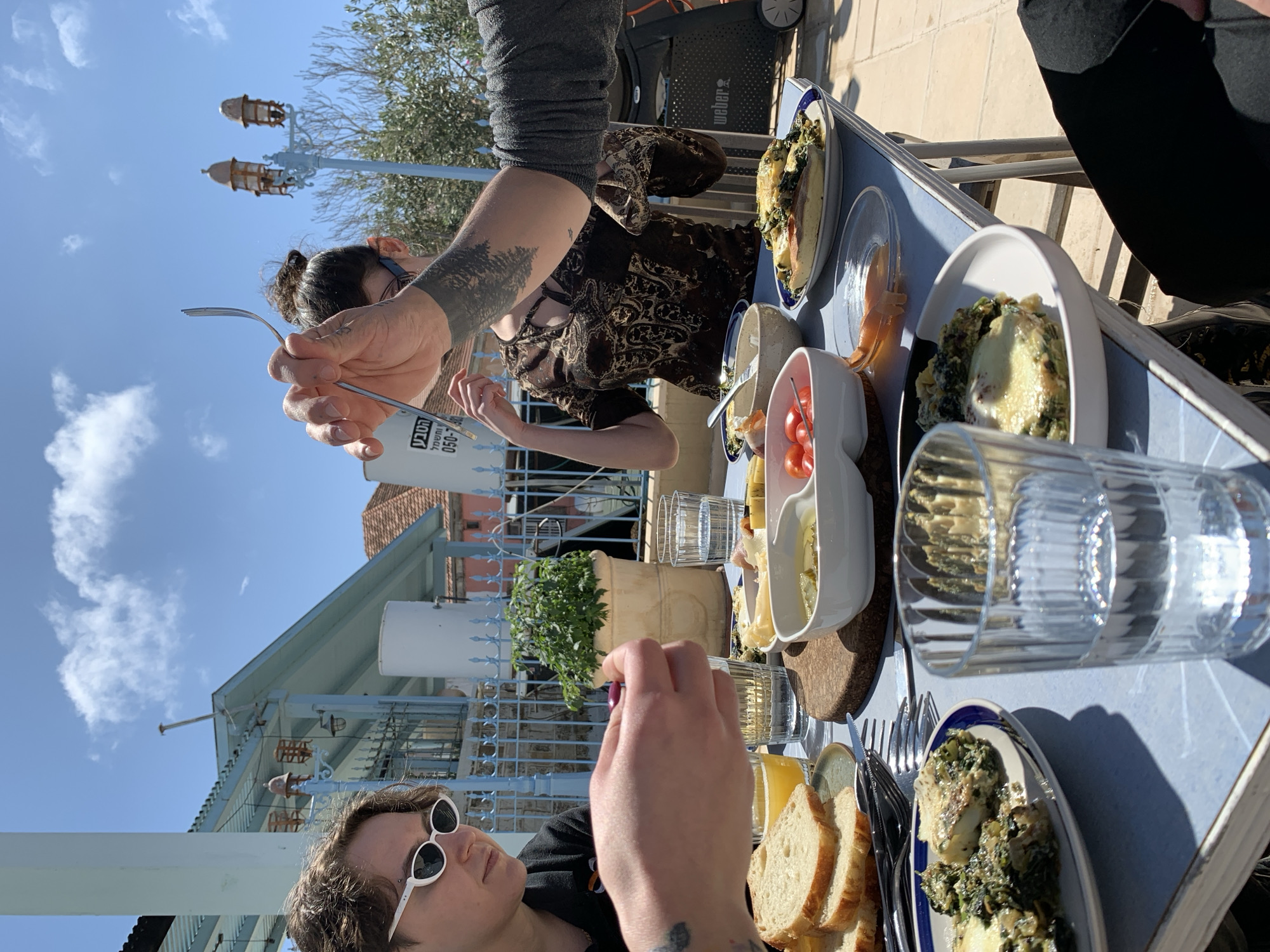 The image shows a group of people dining outdoors on a sunny day. They are sitting around a blue table laden with plates of food, including what appears to be bread, tomatoes, and some kind of baked dish with spinach. There are also glasses and cutlery on the table. Three people are partially visible in the frame. One woman is wearing sunglasses and a black shirt, another woman has a dark patterned top, and a third person has a grey sleeve. A fork is being held up near the woman with her hair up.
In the background, there is a building with a blue and white exterior and a wrought-iron fence. There are also trees, street lamps, and a blue sky with some clouds. The presence of blue and white colors and the architecture style is consistent with the city of Tel-Aviv, Israel.