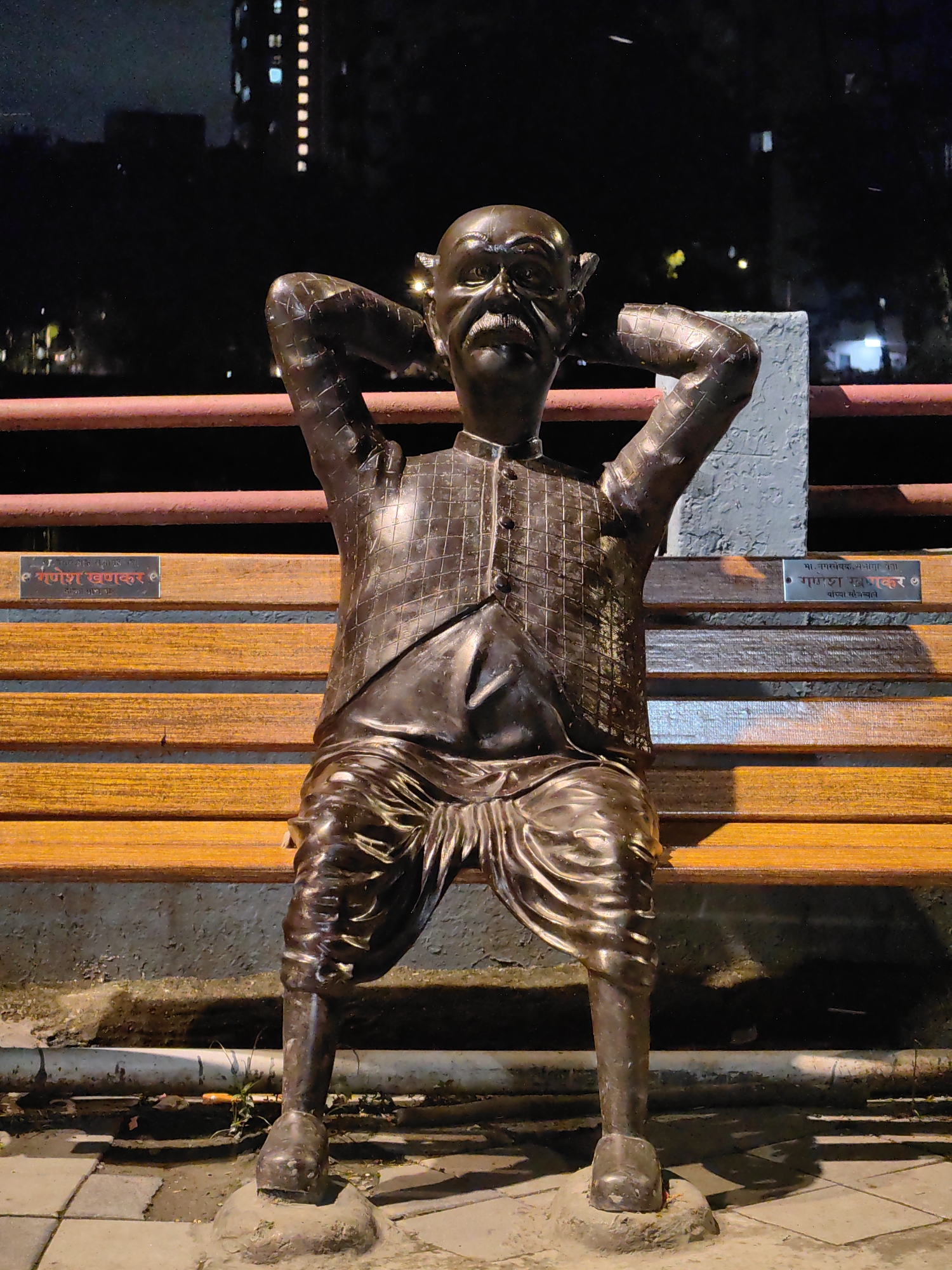 A bronze statue of a man with a mustache is seated on a wooden bench at night. The statue is wearing a traditional Indian outfit, including a vest and pants. The man has his hands behind his head, as if relaxing. Behind the bench, there is a red railing and a stone wall. In the background, a tall building with illuminated windows is visible. There are two metal plaques on the bench, one on the left and one on the right, with inscriptions in an Indian language. This statue is located at the Bandstand promenade in Mumbai, India.