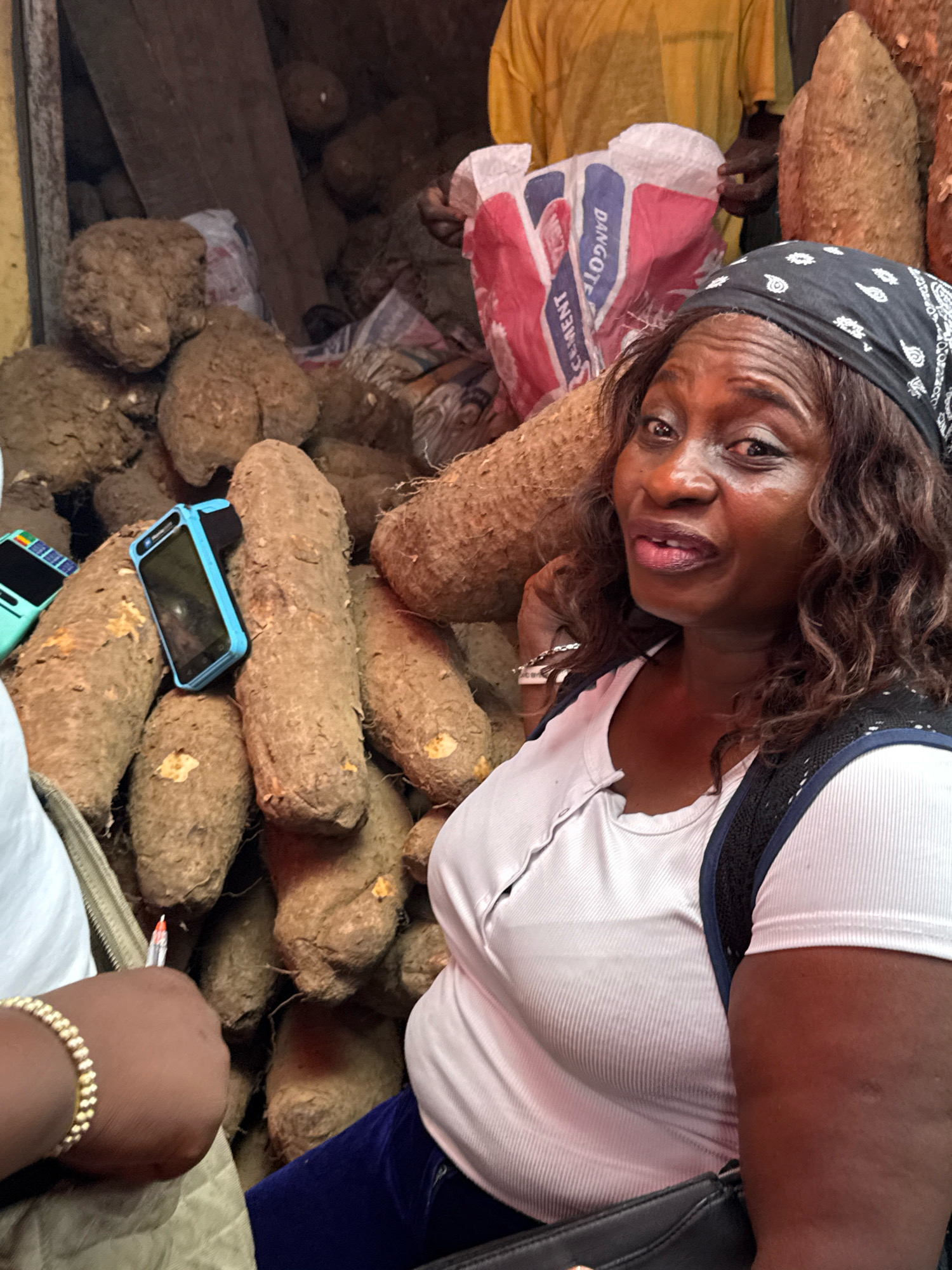 A medium shot captures a woman from Nigeria, possibly at a market in Ajah, Nigeria, surrounded by large yams. The woman, wearing a black bandana and a white ribbed t-shirt, gazes towards the viewer with a slightly surprised expression. A black backpack is slung over her shoulders. To her left, a blue card reader is placed on a yam. In the background, a yellow shirt is visible, along with a red and white bag labeled "Dangote Cement." The scene is likely a vibrant market setting given the abundance of produce and the commercial packaging.