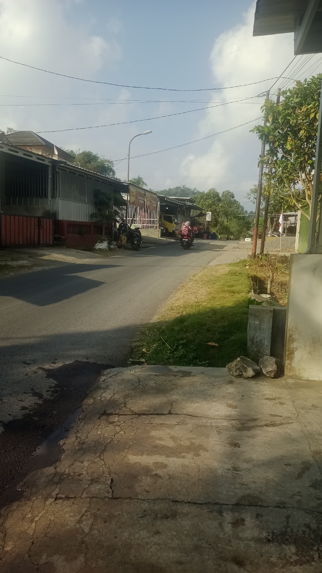 The image depicts a street scene in Tonjong, Indonesia, on a clear day with scattered clouds. The foreground shows a cracked concrete pathway with puddles of water on the adjacent asphalt road. A paved road curves to the left, lined with buildings featuring corrugated metal roofs and some with red and gray siding. There are parked motorcycles and a yellow vehicle visible. Further down the road, a person wearing a red hijab is riding a motorcycle away from the camera, and another motorcycle is parked near a building. Trees and foliage are present on both sides of the road, and utility poles with overhead wires are visible. A street lamp stands on the left side of the road. Two large rocks are placed near a concrete structure in the lower right portion of the image.