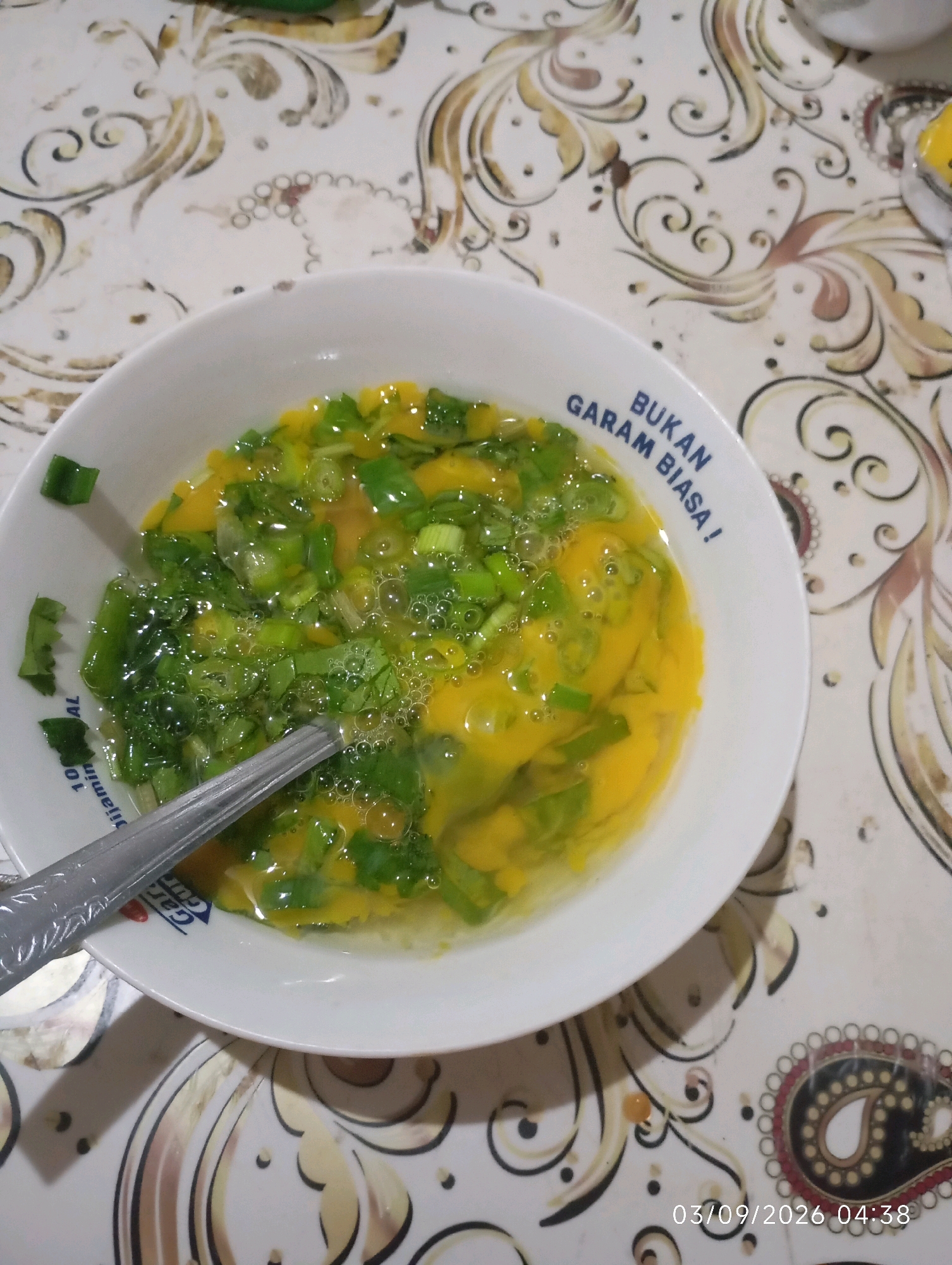 A close-up, eye-level shot captures a white bowl filled with a mixture of beaten eggs and chopped green onions, appearing to be ready for cooking. The bowl rests on a patterned tablecloth with ornate swirls in cream and gold. A silver spoon rests partially submerged in the egg mixture. Text is visible on the rim of the bowl in blue lettering that reads, "BUKAN GARAM BIASA!". In the lower right corner of the image, a timestamp indicates "03/09/2026 04:38". The scene is likely from a home kitchen in Klecorejo, Indonesia.
