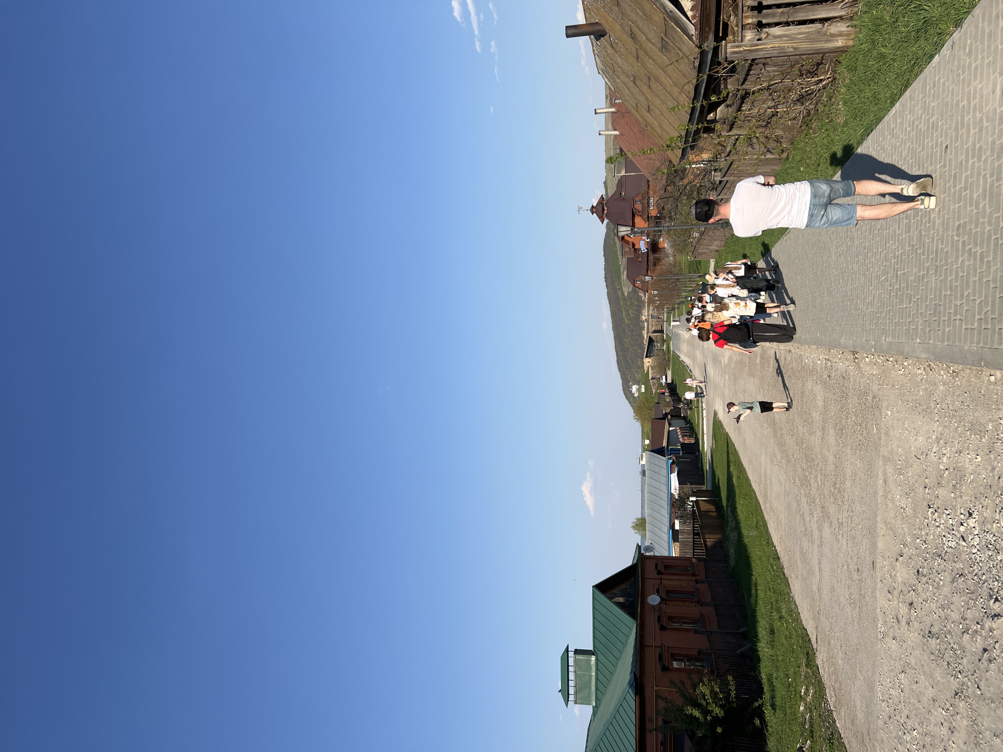 The image shows a street scene in Sviyazhsk, Russia. The view extends along a pedestrian walkway, constructed from light-colored paving stones on the right side and a packed gravel path on the left.
Several people are visible on the walkway, including a man in a white shirt and blue shorts near the top right, casting a shadow on the paving stones. Further down the path, there is a group of people. On the lower section, a person is lying on the gravel path.
On both sides of the walkway, buildings of various sizes and architectural styles can be seen, with some having wooden exteriors and traditional designs. Patches of grass grow between the buildings and the walkway.
The sky is a clear, deep blue and takes up a large portion of the left side of the frame.