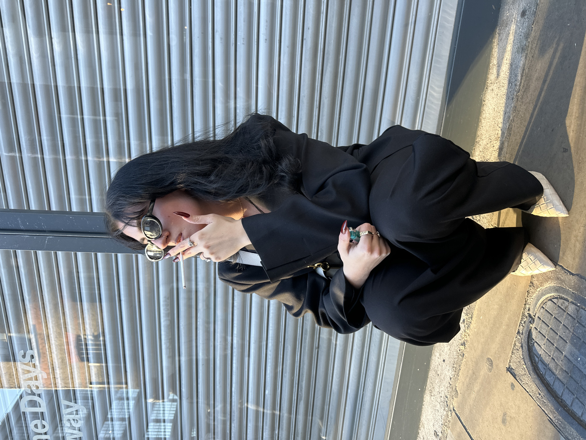 A person is captured in a crouched or squatting position on a sunny urban sidewalk in London, United Kingdom. The individual, with long dark hair, is wearing oversized dark sunglasses, a black long-sleeved top or jacket, and black wide-leg trousers. They have dark red nail polish and wear multiple rings on their fingers, including one with a prominent green stone. A lit cigarette is held to their mouth, indicating they are smoking. Their gaze is directed towards the viewer's right.
Behind the person is a grey corrugated metal roller shutter, likely belonging to a closed shop or establishment. Reflected on what appears to be glass in front of the shutter, partially obscured and rotated, is some faint text including "one Days" and "Way". The ground is a mix of concrete and asphalt, showing strong shadows cast by the person, suggesting it is daytime with a low sun angle. A circular metal manhole cover with a grid pattern is visible on the pavement near their white, textured flat shoes.