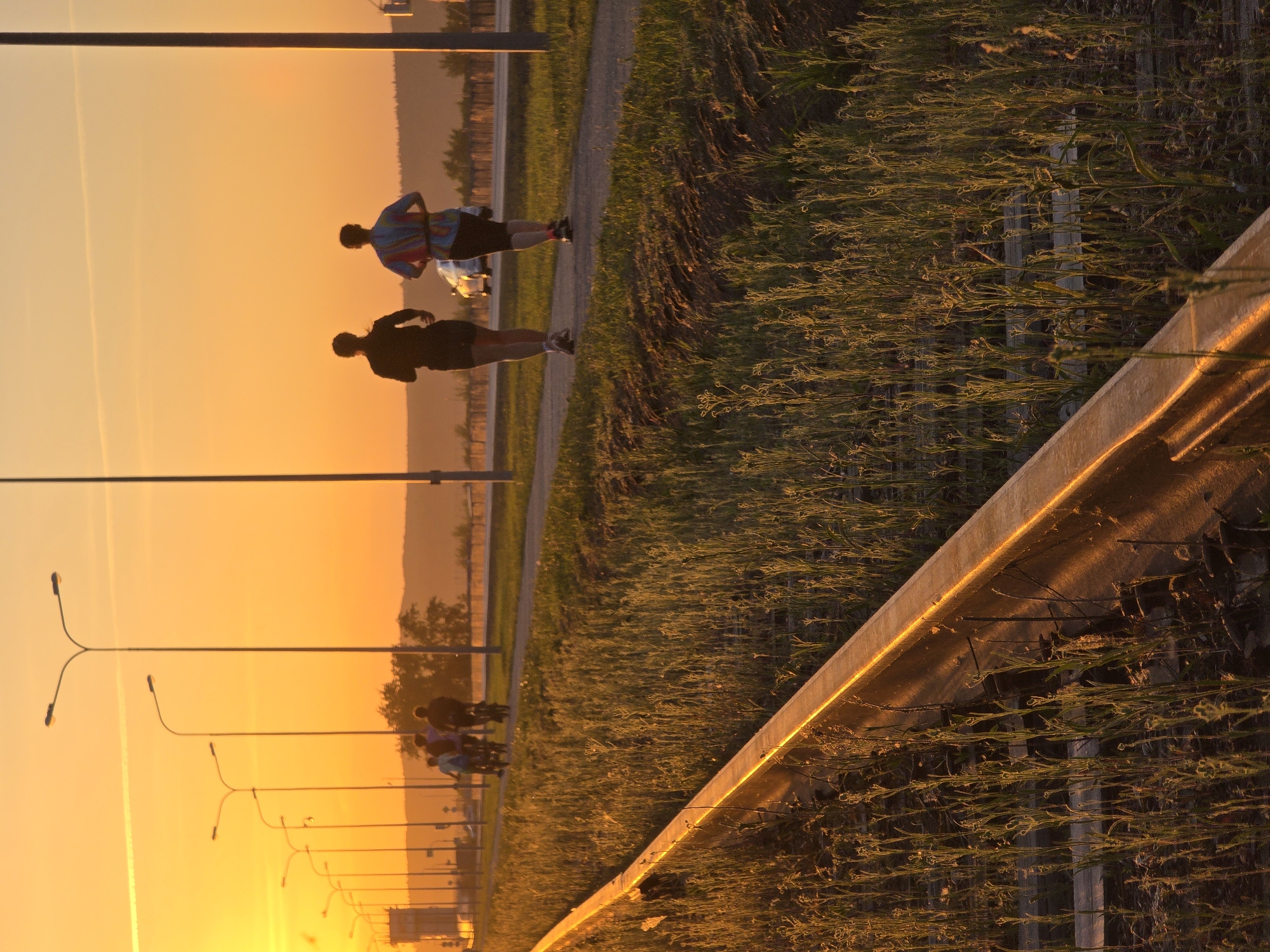 A horizontally rotated image captures a scene at sunrise or sunset, likely in Chaikovsky, Russia, given the request. Two individuals are running on a paved path alongside a grassy area and a road with street lights. One runner is wearing a colored shirt, while the other is in a dark outfit. The scene is bathed in warm, golden light. A concrete barrier runs diagonally across the foreground. The sky is orange.