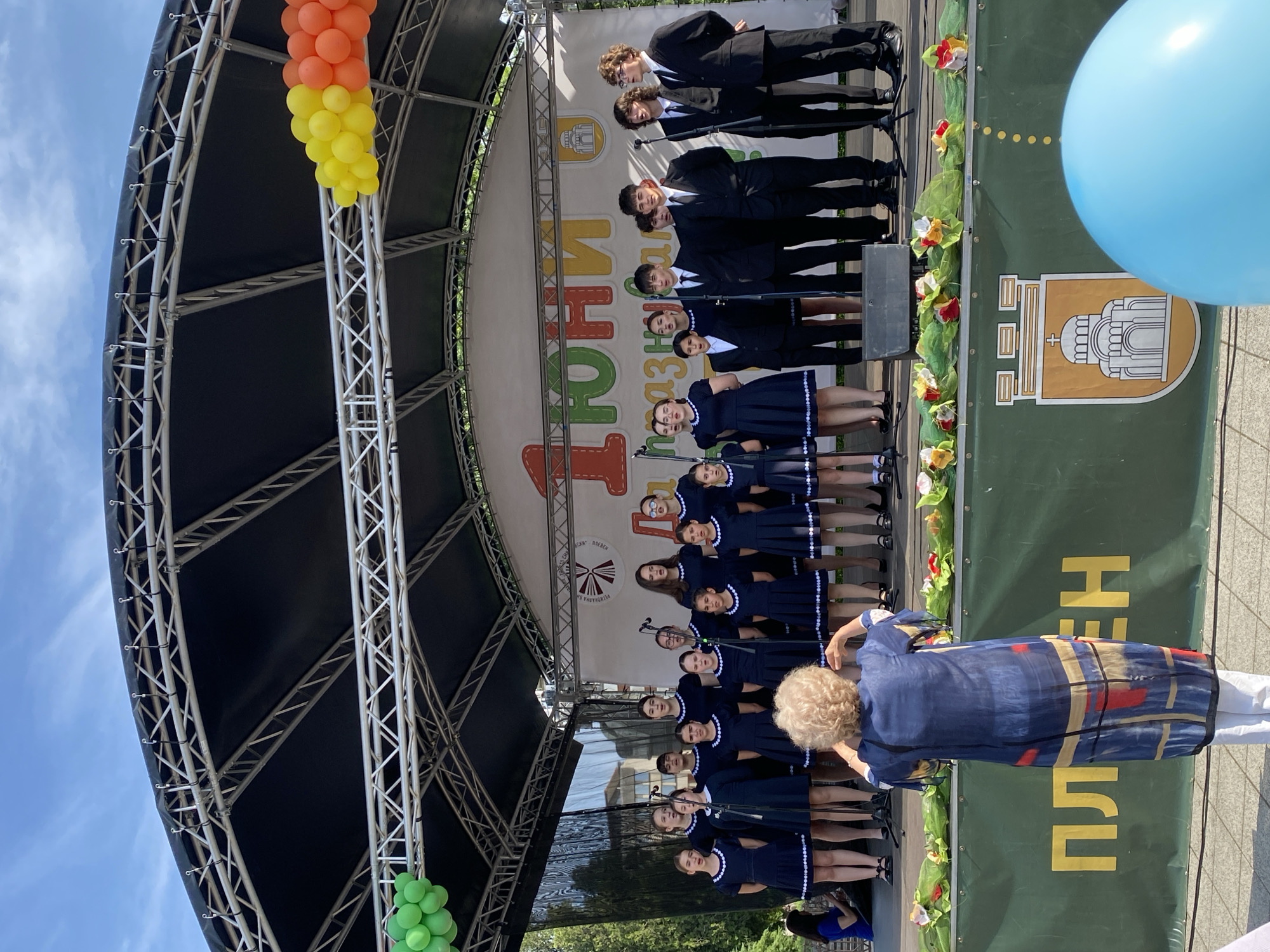 A low-angle shot depicts a youth choir performing on an outdoor stage in Pleven, Bulgaria. The choir consists of approximately 20 girls in matching navy blue dresses with white trim and five boys in dark suits. They stand in rows, singing into microphones on stands. The stage is covered by a metal truss structure with a dark canopy. Balloons in orange, yellow, and green are attached to the truss. A large green banner with a stylized building and writing is visible behind the choir. A woman in a blue dress with geometric patterns stands in front of the choir, possibly directing or photographing them. A blue balloon floats to the right of the stage. The sky is partially visible and blue.