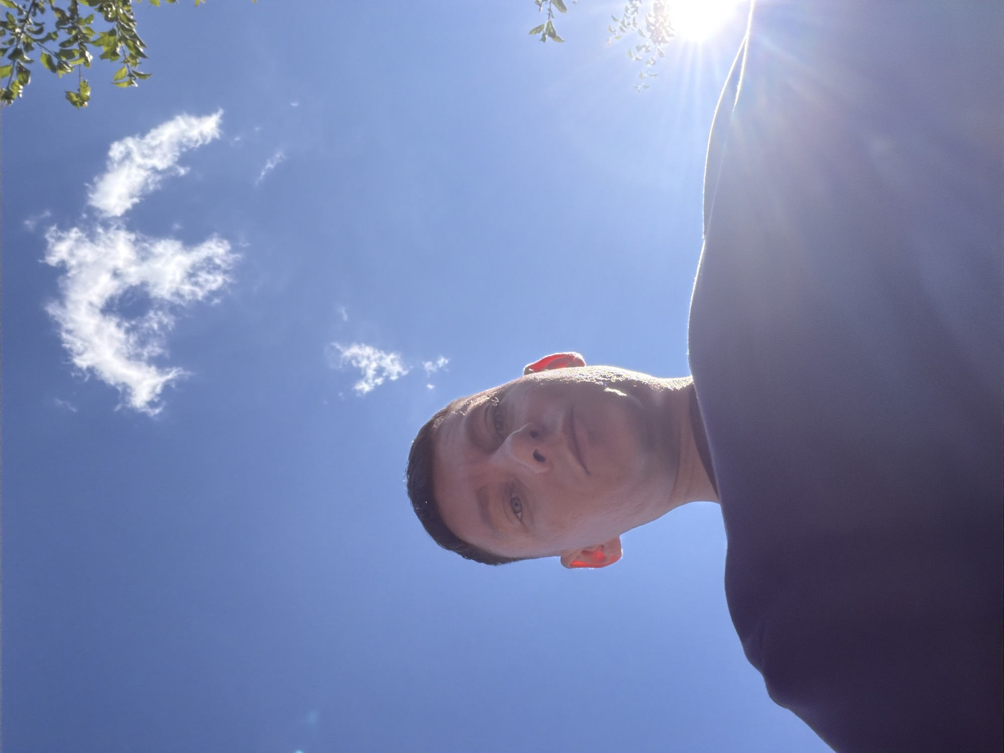 A low-angle, close-up, outdoor, waist-up shot shows a person with short dark hair and fair skin from the chest up, facing upwards towards the sky. The person's face is slightly illuminated from the right, with a serious expression. Their eyes are open and looking directly at the camera. The person is wearing a dark-colored garment, possibly a shirt or sweatshirt. The background consists of a clear blue sky with a few wispy white clouds. A bright sun is visible in the upper right quadrant, casting lens flare and highlighting the edge of the person's face and garment. Some green leaves from a tree are visible in the upper right corner, silhouetted against the sky. The image appears to have been taken during daylight hours.
This image does not contain any information that can be used to determine its location in Moscow, Russia.