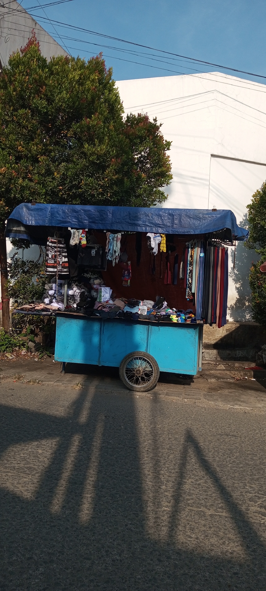 A mobile clothing stall is set up on the side of a street in Setu, Indonesia. The stall is covered by a blue tarp and displays various garments, including shorts, socks, underwear, and pants, hanging on a rack or folded on shelves. The stall is mounted on a blue cart with a single spoked wheel. In the background, there is lush green foliage, a white building, and a clear blue sky. The ground in front of the stall is paved with asphalt, and the shadows of the stall and trees are cast across it.