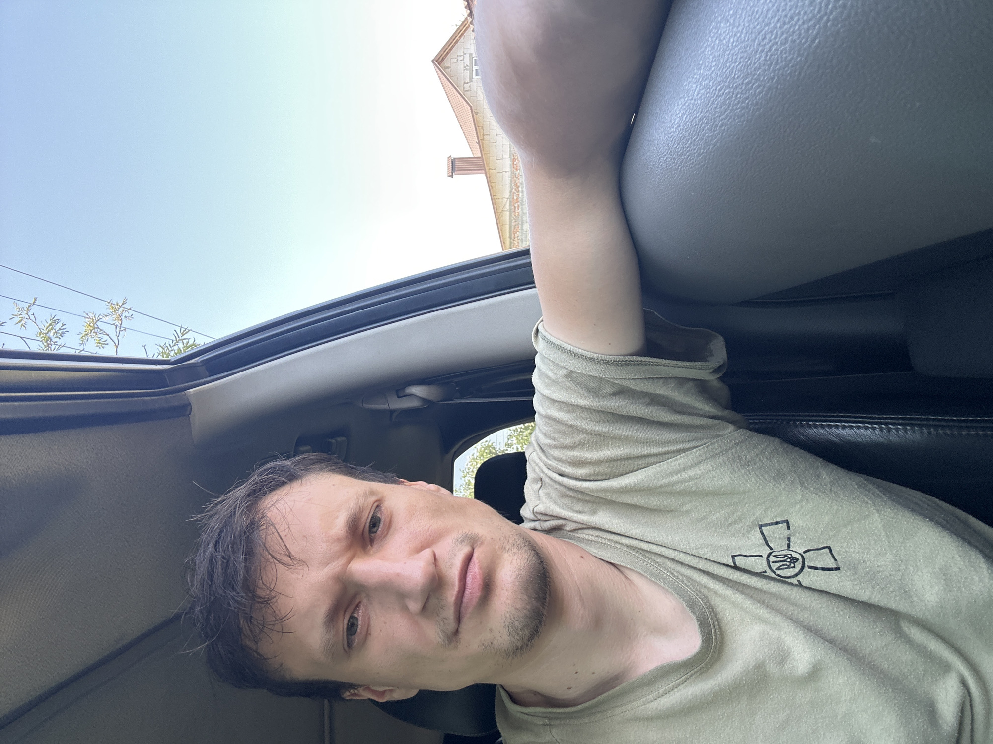 A man with brown hair and a light complexion is sitting in the driver's seat of a vehicle. He is wearing a light green t-shirt with a black emblem on the left chest. His facial expression is neutral. The interior of the vehicle is visible, including the dashboard and part of the passenger seat. Outside the car, a clear blue sky can be seen, with some trees and a building with a tiled roof in the distance. The building appears to be a house in Makiv, Ukraine.