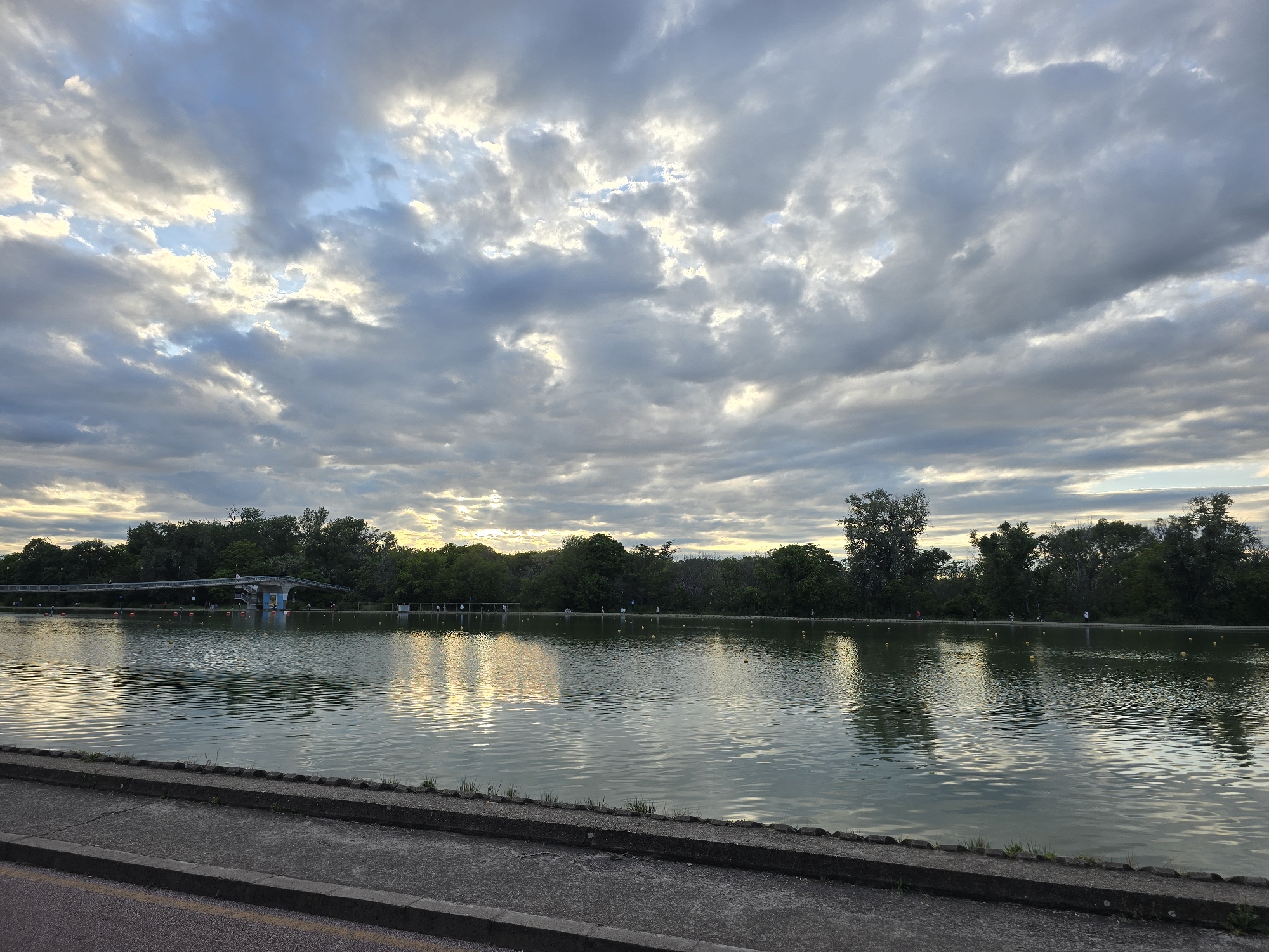 The image depicts a body of water reflecting a cloudy sky. In the background, a line of trees and a structure resembling a pedestrian bridge are visible. Yellow buoys are scattered across the water. In the foreground, there is a concrete edge and a portion of an asphalt surface. The city and specific location are Unknown.