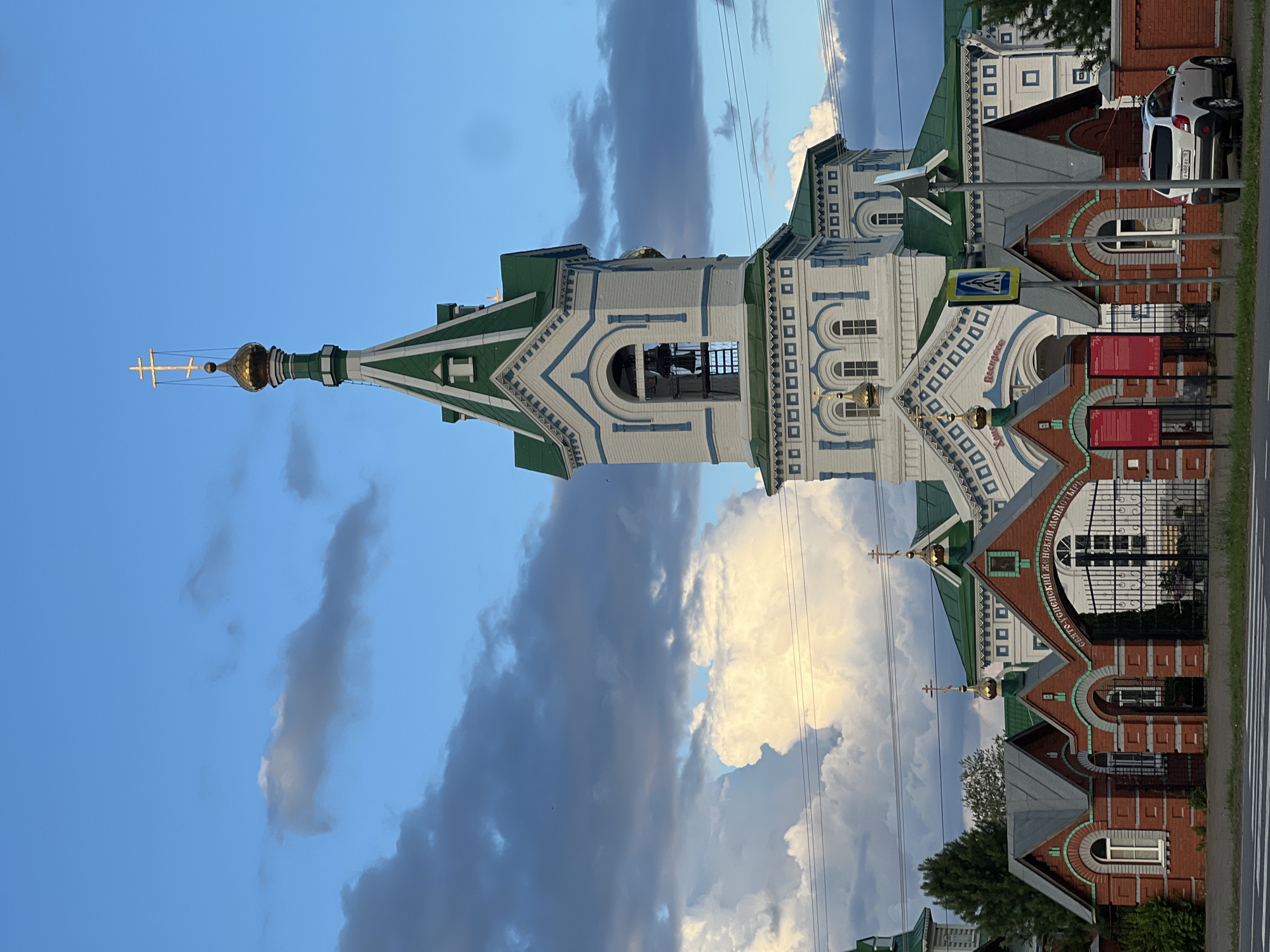 The image depicts a vertical shot of an orthodox church with a white facade and green roof, set against a backdrop of a partly cloudy blue sky. The church features intricate architectural details, including decorative white patterns against a blue border on the white walls, arched windows, and several golden domes topped with crosses. A prominent bell tower is visible, with an open belfry. The church is situated next to a road, and a white car is parked alongside it. The lighting suggests it is either early morning or late afternoon, with the sun illuminating some of the clouds and highlighting the golden domes. The overall mood is serene, with the majestic architecture of the church dominating the frame. Visible text on the building reads "ВОСКРЕСЕНСКИЙ ЖЕНСКИЙ МОНАСТЫРЬ", which translates to "Resurrection Women's Monastery". The location is identified as Perevoznoye, Russia.