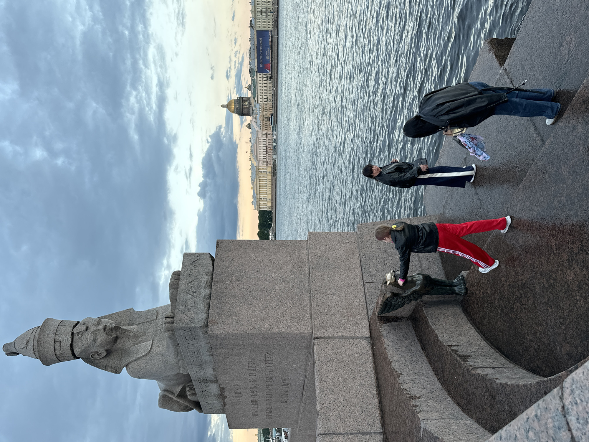 This image depicts the Sphinxes on the University Embankment in Saint Petersburg, Russia, at dusk. A large granite sphinx statue dominates the left side of the frame, with hieroglyphs carved into its base. In the foreground, three individuals are visible. A young girl in a black jacket and red pants with white stripes on the side is touching a small winged creature sculpture at the base of the sphinx. To her right, a woman in a black jacket and dark blue skirt stands facing the water. Further to the right, another person in a black jacket and blue jeans is bending down, looking at something in their hands. The Neva River is in the background, with a building complex on the opposite bank, topped by a golden dome, likely the Admiralty Building. A blue banner with Russian text is visible on the building. The sky is overcast with dramatic clouds. The overall atmosphere suggests a cool evening in the city.