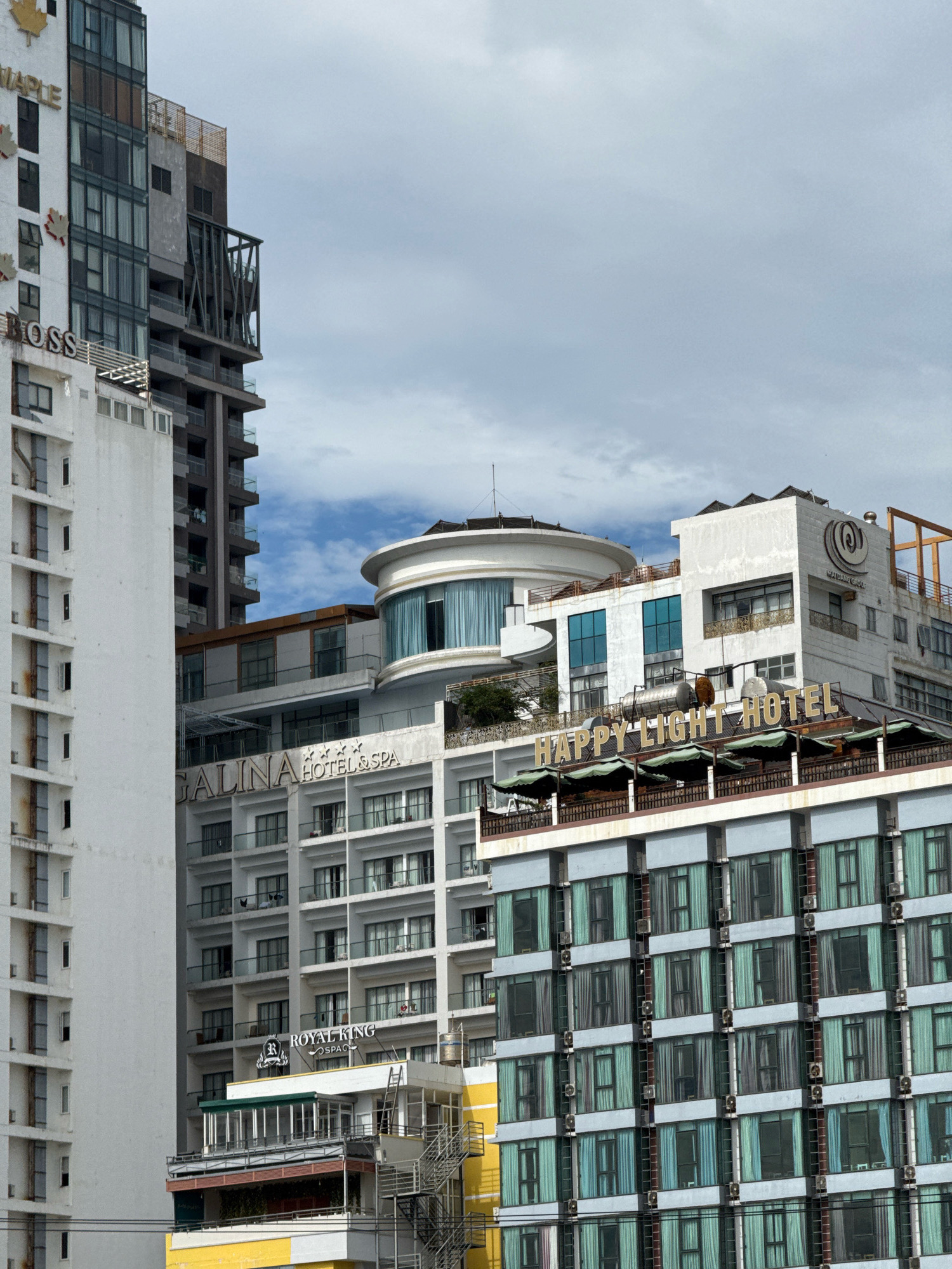 The image shows a cluster of multi-story buildings in Nha Trang, Vietnam. The foreground features a tall, white building with rows of balconies and windows, some appearing slightly weathered. To its right, a white building with the sign "GALINA HOTEL & SPA" is visible, featuring a modern architectural design with balconies and windows. Further to the right and slightly behind, a building with the prominent sign "HAPPY LIGHT HOTEL" is depicted, also with multiple floors of windows and balconies. Above these, a circular building with large windows and a rooftop structure is present. The sky above is partly cloudy. The buildings exhibit a variety of architectural styles, common in urban environments. There are no visible people or specific actions occurring in the frame.