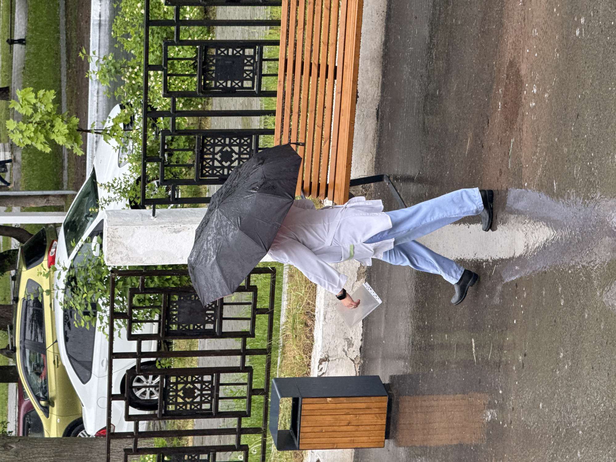 A person in light blue trousers, a white lab coat, and black shoes walks in the rain holding a black umbrella and a plastic folder in Chaikovsky, Russia. The person is walking on wet pavement. A wooden and black metal bench and an accompanying waste container are near a black metal fence. Behind the fence are parked cars, one yellow and one white. Trees and grass are also visible behind the fence.