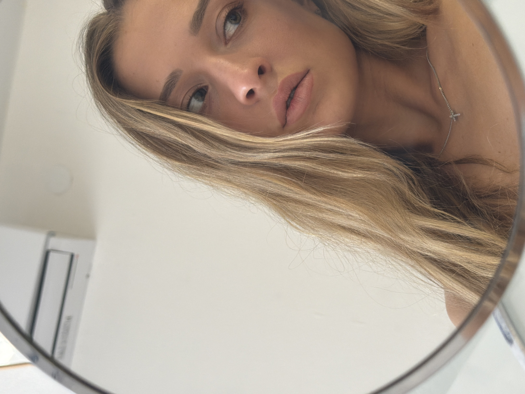 The image presents a close-up, angled view of a woman's face reflected in a round mirror. She has light skin, blonde hair with darker roots, and light-colored eyes. Her gaze is directed upwards and to the left. She is wearing a delicate silver necklace with a small cross pendant. The background is a plain white wall with a small white object visible to the left of the mirror. The image does not show any indications of being in Khimki, Russia, or any details allowing for the confirmation of it being taken there.