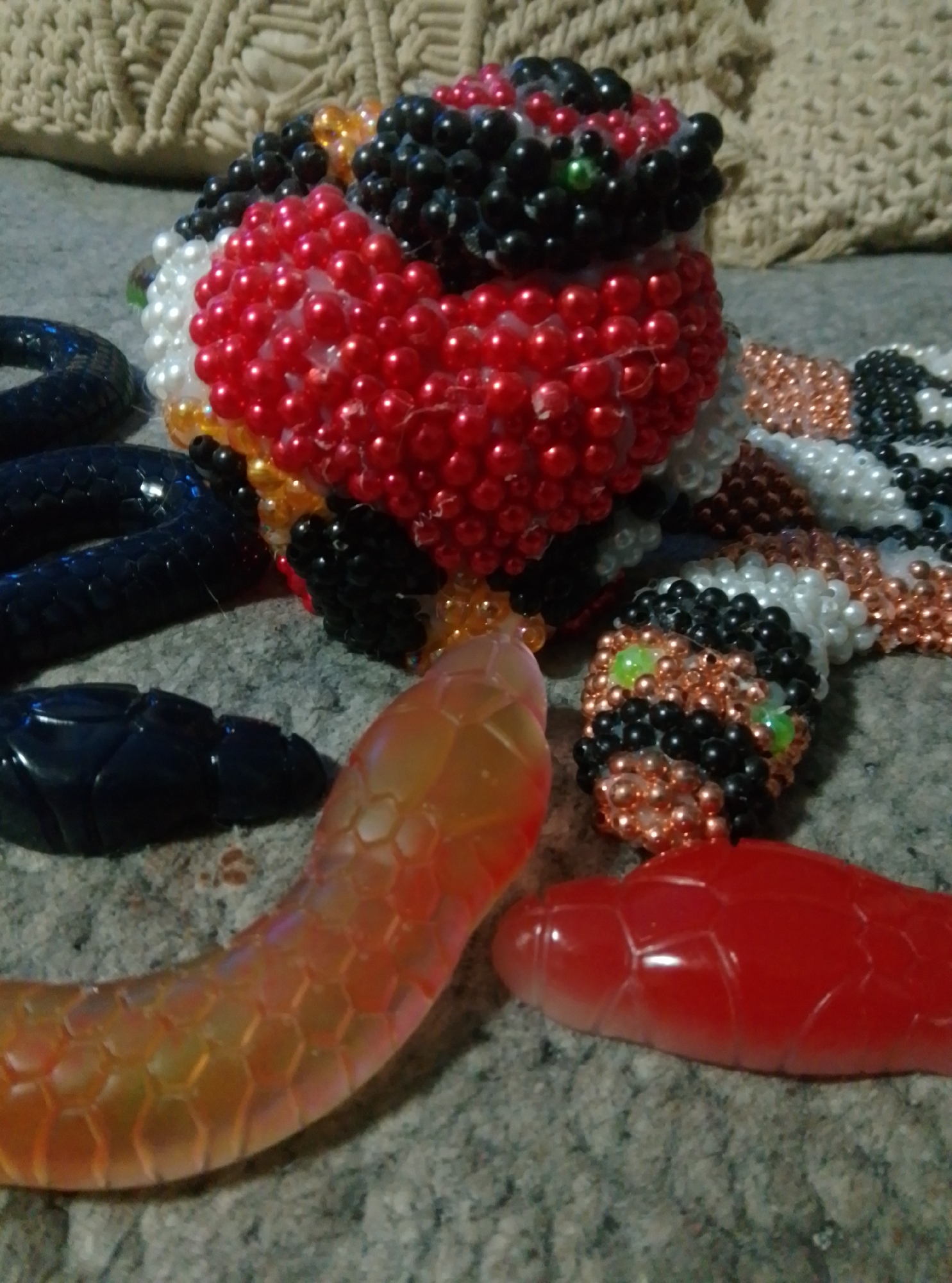 The image depicts a close-up, eye-level view of several snake-like objects and a beaded heart, arranged on a textured surface that appears to be a carpet or rug. The objects are not located in the city of Wedel, Germany; this is a generic indoor scene.
Dominating the foreground are two translucent, segmented objects resembling plastic snakes or toys. The left object is a vibrant orange-yellow, with a hexagonal scale pattern visible beneath its surface. The right object is a solid red, also featuring a segmented, scale-like texture.
Behind these, a larger, intricately crafted object made of small beads is prominent. It forms a heart shape at the front, primarily composed of red and white beads, with accents of black and clear beads. Extending from the heart is a beaded snake body, featuring segments of black, white, and copper-colored beads. The head of this beaded snake is visible on the right side of the image, with black beads forming its outline and two green beads serving as eyes. There are also sections of dark blue, segmented plastic that resemble snake bodies scattered across the scene.
In the background, a textured, woven material in a neutral color, possibly a blanket or upholstery, is visible. The overall lighting suggests an indoor environment, with diffused light sources. The composition is a still life arrangement of decorative objects.