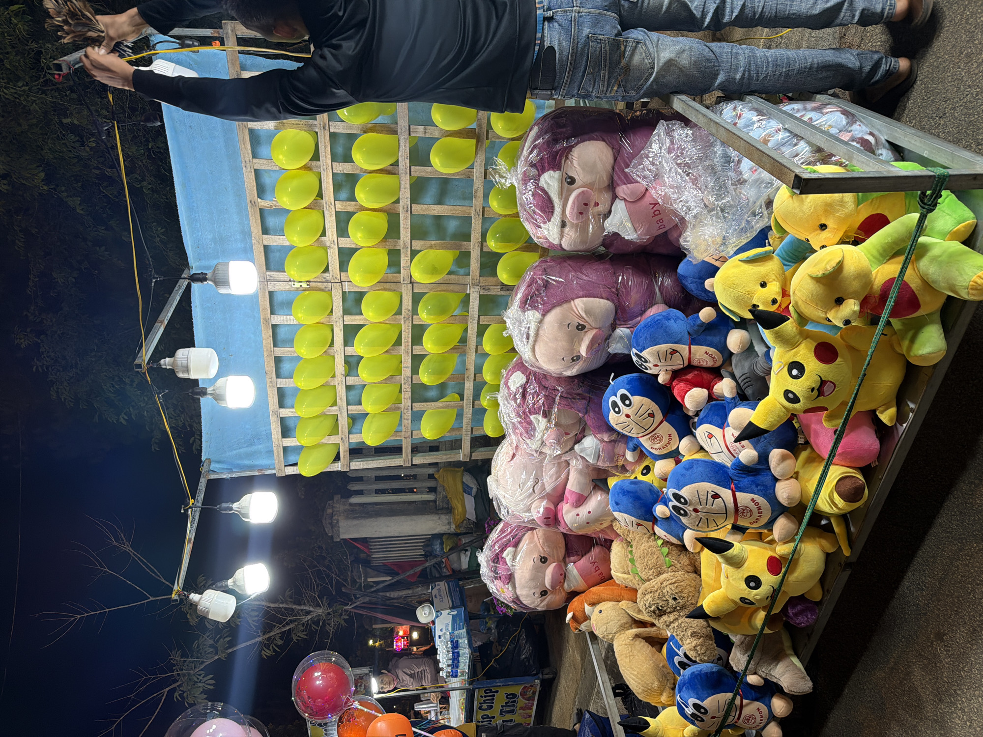 This image captures a vibrant night scene at an outdoor market or fair in Măng Đen, Vietnam.
**Subjects:** The primary human subject is a man, visible from the waist up, wearing a black long-sleeved shirt and jeans, focused on aiming a dart gun or similar device. A second person is partially visible at a distant food stall. The main objects include a wall of yellow balloons set within a wooden lattice, numerous bright LED light bulbs illuminating the scene, and a large display of plush toys acting as prizes. The plush toys consist of purple pig-like characters (some labeled "baby"), blue Doraemon figures, yellow Pikachu figures, and other assorted animal or character plushies, many wrapped in clear plastic bags. In the background, there is a food stall with bottled water and a sign. Also visible are decorative inflatable balloons with internal lights (red, orange, pink) in the lower left.
**Activity or event taking place:** The man is engaged in a classic carnival game, aiming to pop balloons, likely to win one of the many plush toy prizes. This activity is part of a larger night market or fair event.
**Notable details:** The image is taken at night, with the dark sky contrasting against the bright artificial illumination provided by the LED bulbs. The weather appears clear, suitable for an outdoor event. The atmosphere is lively and engaging, typical of entertainment venues. Many of the yellow balloons on the wall are deflated, suggesting active gameplay. The general setup, with game stalls and prizes, provides a strong location cue for a fair or market.
**Any visible text:** Recognizable text includes "baby" printed on some plush toys, "DORAEMON" on the blue plushies, "KEMON" (likely part of "POKEMON") on some yellow plushies, "AQUARINA" on water bottles at the background stall, and "Tip Chip Thơ" on a sign.