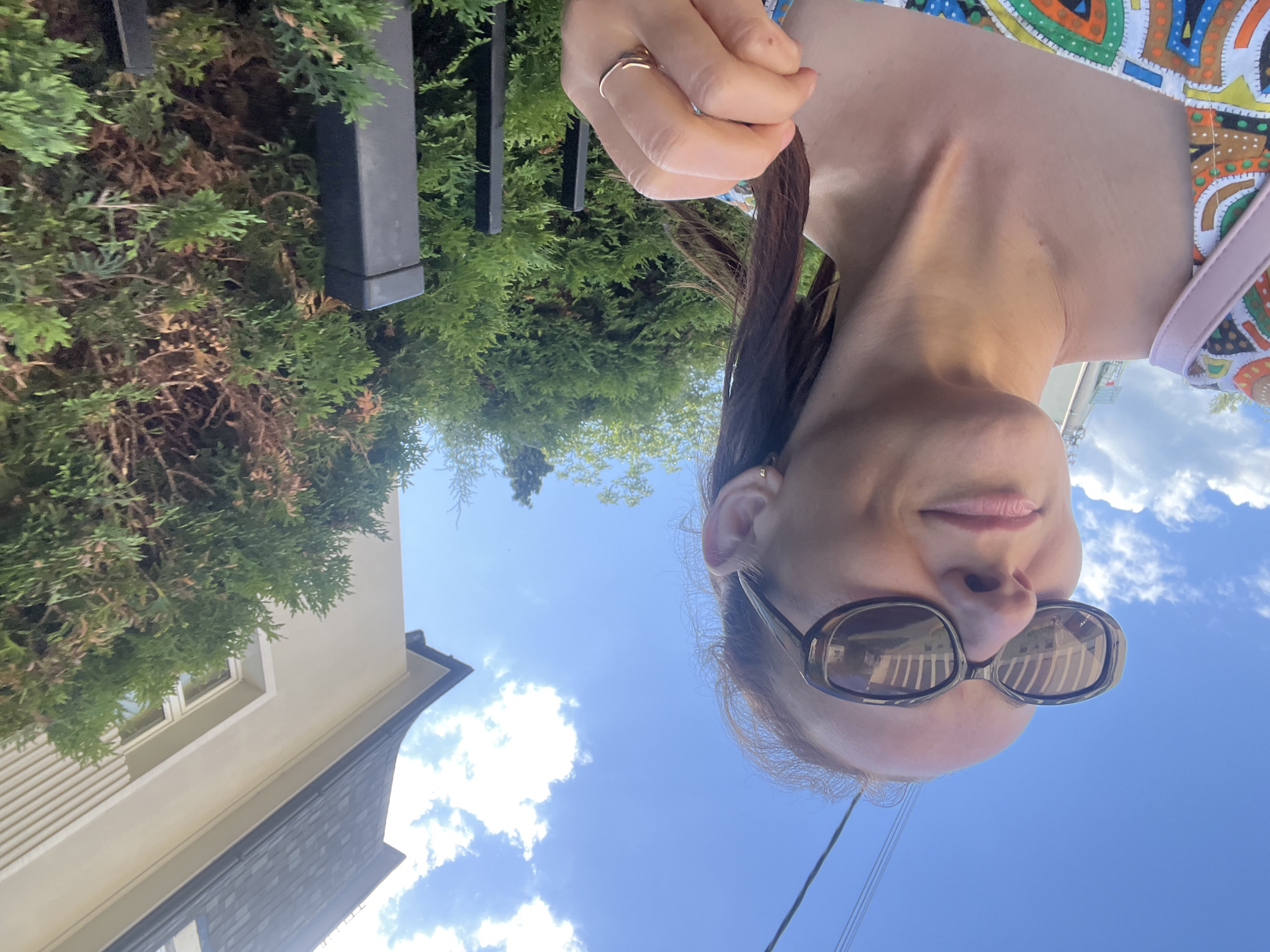 A close-up, eye-level outdoor selfie shows a woman wearing sunglasses and a colorful dress. She is looking towards the left of the frame, with her face partially illuminated by sunlight. Her hair is pulled back, and she is wearing a gold ring on her right hand. In the background, a building with a dark, mansard-style roof is visible, along with green trees and a bright blue sky with some clouds. The scene appears to be in a residential area.