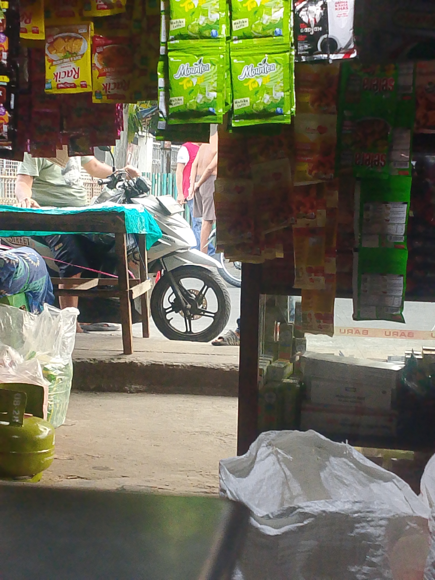 The image depicts a street-side stall in Palembang, Indonesia, characterized by an array of packaged goods hanging from the ceiling and shelves. In the foreground, a green LPG gas cylinder with markings "08-24" is visible, alongside plastic bags filled with merchandise. A wooden table, draped with a turquoise cloth, is situated in the center, partially obscuring a silver motorcycle. A person, wearing a light-colored t-shirt and dark pants, is seated behind the table, seemingly attending to the stall. Further back, two individuals are standing, one in a red t-shirt and another in a light-colored shirt and shorts, suggesting a commercial transaction or social interaction. The environment appears to be an open-air market or shop, with concrete flooring and a slight elevation leading to the stall area. The overall impression is one of a typical local retail setting within a bustling urban environment of Palembang.