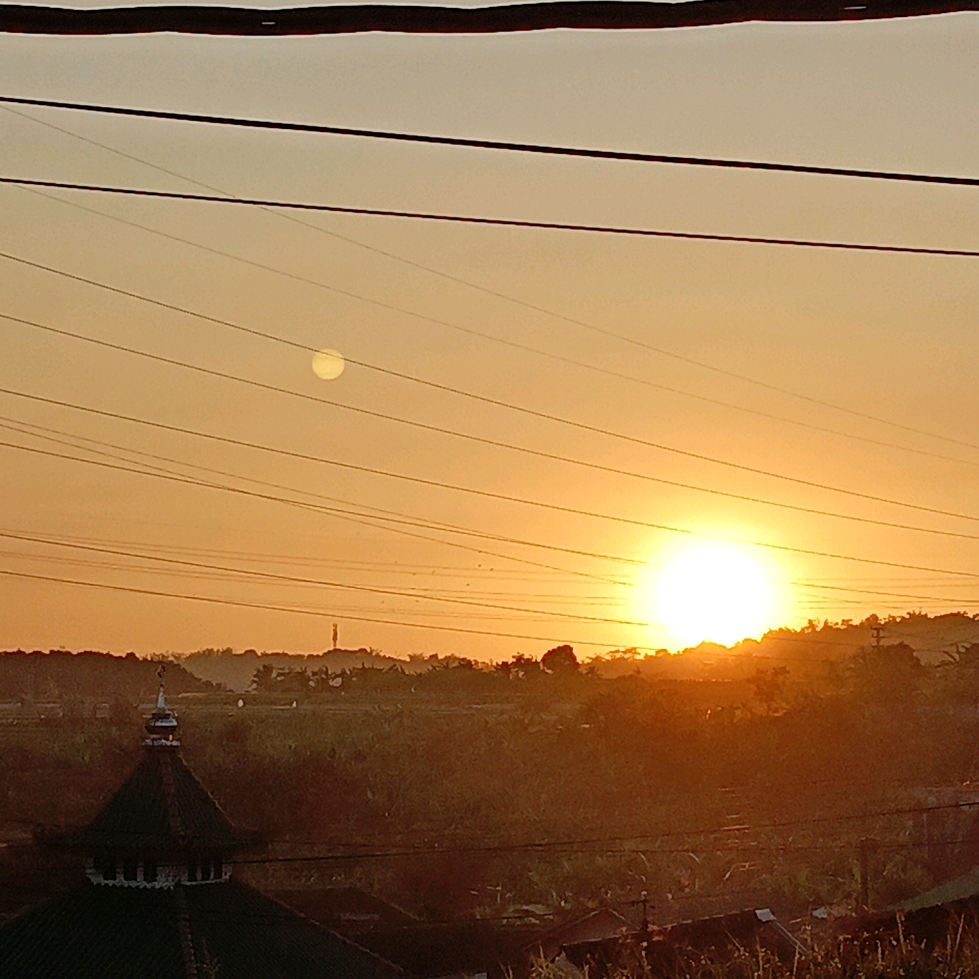 The image depicts a sunset scene in Salatiga, Indonesia, characterized by a hazy orange sky and silhouetted power lines stretching across the frame. In the distance, a glowing sun is prominently visible on the right side, with a smaller, less distinct sun or moon appearing higher in the sky on the left. The foreground features the dark silhouette of a building with a dome and minaret, likely a mosque, on the left side of the image. The mid-ground shows a dense, dark landscape of trees and vegetation, possibly indicating a hilly terrain. Various power poles and wires are scattered throughout the scene, suggesting an urban or semi-urban environment. The overall impression is one of a warm, serene sunset over a landscape marked by both natural elements and man-made infrastructure.