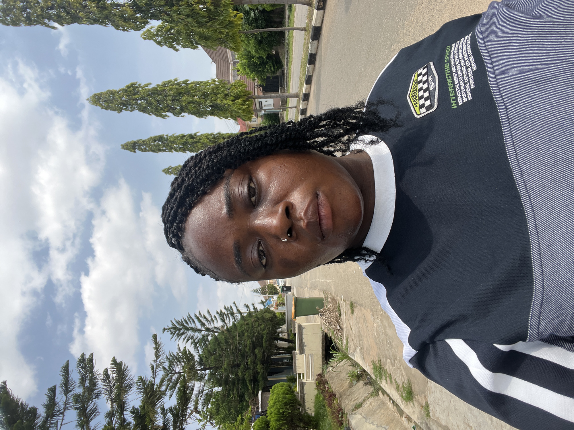 In the city of Abuja, Nigeria, a medium-shot, eye-level selfie captures a young Black woman with braided hair and a nose ring standing on a paved residential street. She is looking directly at the camera with a neutral expression. The woman is wearing a black long-sleeved shirt with white stripes on the shoulders and sleeves, and a graphic design on the left chest.
The background reveals a street lined with tall, slender trees and manicured bushes. On the left side of the street, a drainage channel runs alongside a paved walkway. Further back, residential buildings with brown roofs are visible, interspersed with more trees and vegetation. A green trash bin is positioned on the side of the street, near the drainage channel. The sky above is partly cloudy, with a mix of white clouds and blue sky. The lighting suggests it is daytime, likely a sunny afternoon. The overall scene depicts a typical urban residential environment in Abuja.