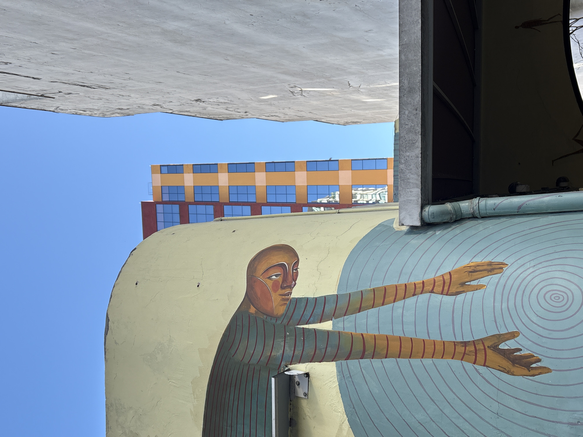 The image depicts a section of a mural painted on a curved, pale yellow wall in São Paulo, Brazil. The mural features a stylized human figure with an orange face and body, rendered with red horizontal stripes on its limbs. The figure appears to be bending forward, with its arms hanging downwards. The background of the mural consists of concentric blue circles.
To the right of the mural, a stark, textured white building wall dominates the frame, creating a strong contrast. In the distance, between the mural and the white building, a modern, colorful building with a grid of orange and red sections and blue windows is visible against a clear blue sky. A grey concrete overhang with a downspout extends into the lower portion of the image, casting a shadow over a dark opening that reveals more of the mural, including a smaller, silhouetted figure. The scene appears to be an outdoor urban environment.
