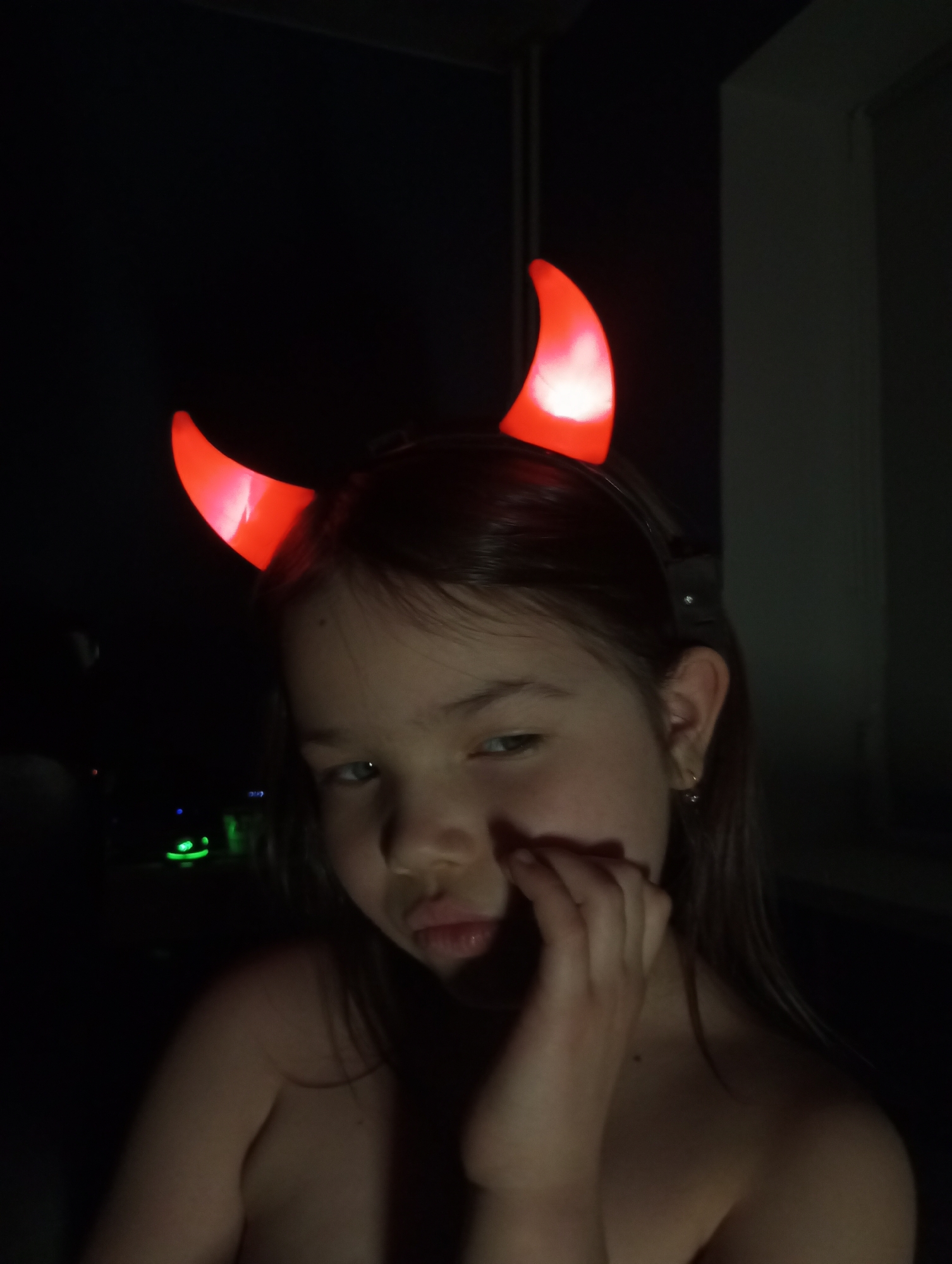 A young girl with dark hair and light skin wears a headband with glowing red devil horns. She is partially visible in a dark environment. Her right hand is raised to her face. There is a light to the left of her, green in color. A light gray wall is visible behind her to the right. There is no visual evidence suggesting the location is Pervomaysky, Russia.