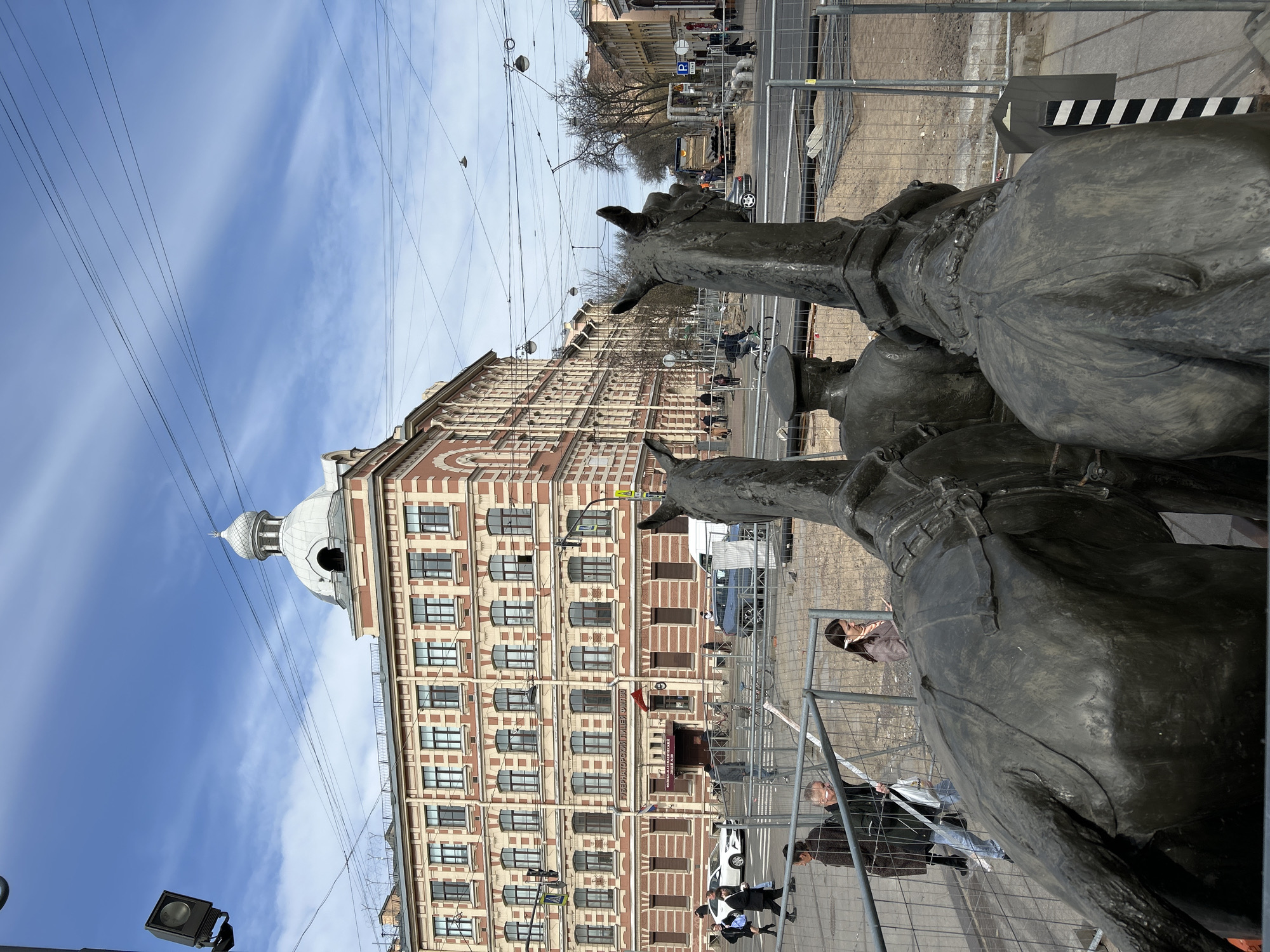 The image presents a daytime urban street scene, likely in Saint Petersburg, Russia, characterized by a mix of historic architecture and contemporary activity. In the immediate right foreground, large, dark bronze statues, appearing to be horse-like figures, are partially visible, situated behind a temporary metal construction fence. Several people are seen walking behind this fence on what appears to be a sidewalk; among them are a man with glasses wearing a dark jacket and a woman in a brown coat.
Dominating the left side of the mid-ground is a grand multi-story building. Its facade features distinctive architectural details, including alternating patterns of light-colored and red brick, numerous arched windows, and ornate trim. A prominent dome-shaped cupola crowns the building's left-hand section. A visible sign on the building displays Russian text, identifying it as "ОБРАЗЦОВАЯ ТИПОГРАФИЯ ИМ. А. А. БОРИСОВА" (Model Printing House named after A. A. Borisov) and "ФЕДЕРАЛЬНОЕ ГОСУДАРСТВЕННОЕ БЮДЖЕТНОЕ УЧРЕЖДЕНИЕ КУЛЬТУРЫ" (Federal State Budgetary Institution of Culture).
A street runs horizontally across the mid-ground, separating the building from the foreground statues and fence. A few vehicles, including a white car and a darker car, are present on the road. Additional metal barriers and what appear to be exposed pipes along the street suggest ongoing construction or maintenance work. Overhead, a network of power lines crisscrosses a bright, clear blue sky dotted with soft, scattered white clouds, indicating good weather conditions. The overall impression is that of an active city environment during a bright day.