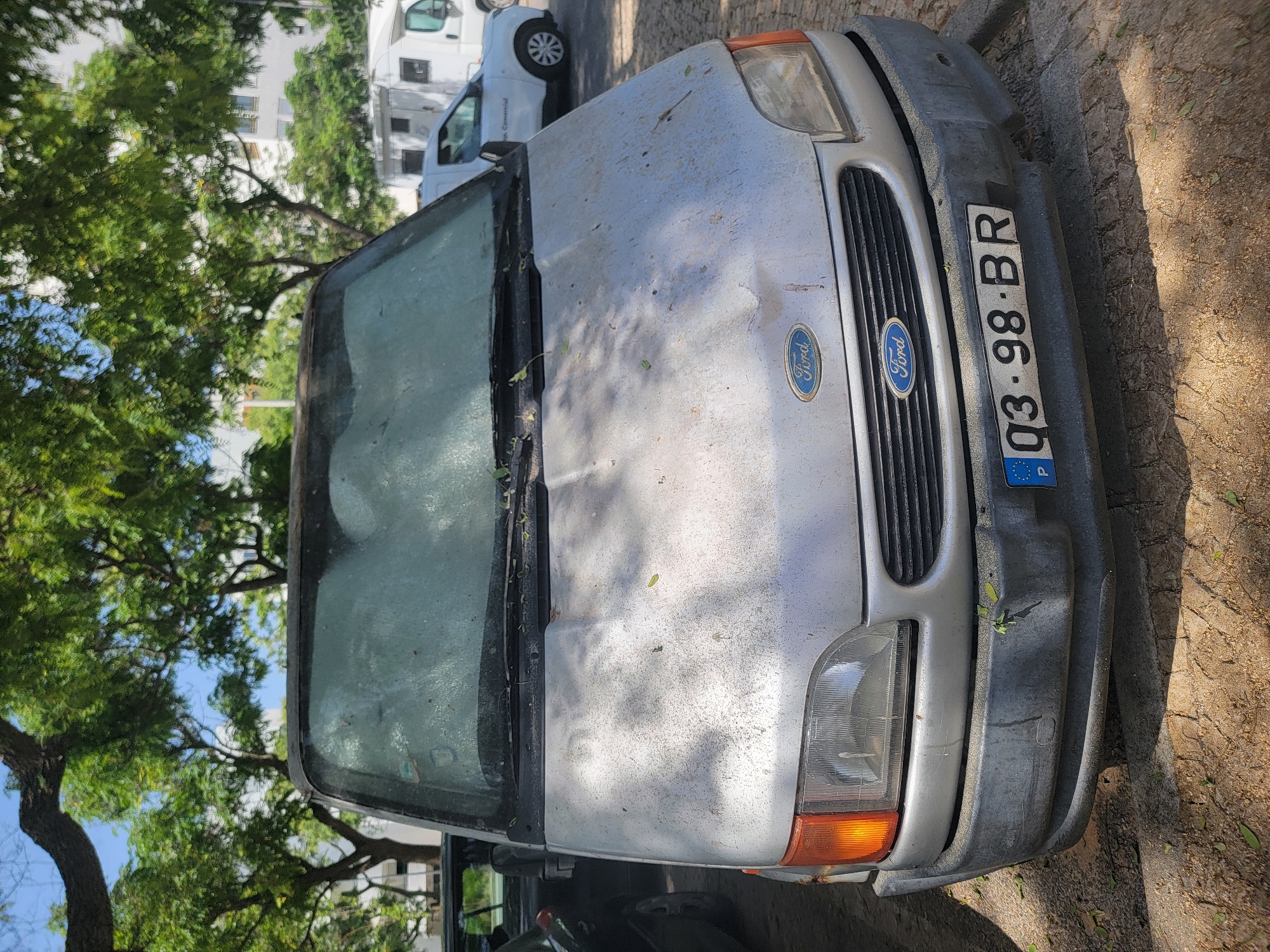 A silver Ford van is parked on a street, angled towards the viewer. The van appears dusty and weathered, with leaves scattered across its hood and windshield. The front of the van is visible, including its grille with the Ford logo, headlights, and an orange turn signal. A license plate with the text "Q3-98-BR" and "P" is attached to the bumper. The van is parked on a street with a cobblestone surface and is partially shaded by trees with green foliage. In the background, a white building and another vehicle can be seen. The scene suggests it is daytime, with natural lighting and shadows cast on the ground. The overall impression is of a van that has been parked for some time.