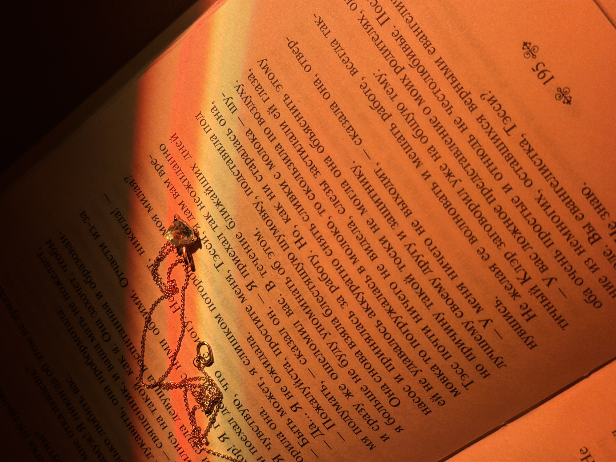 The image shows an open book with text in Russian. A delicate gold necklace with a gemstone pendant lies on the pages, partially illuminated by a warm, concentrated light source, possibly sunlight or artificial lighting, casting a distinct glow on the right side of the book. The page number "195" is visible. The text appears to be part of a narrative or dialogue. The lighting and arrangement suggest a staged or styled photograph, likely for aesthetic or promotional purposes. The location in the city of Chaikovsky, Russia is not discernible from this image.