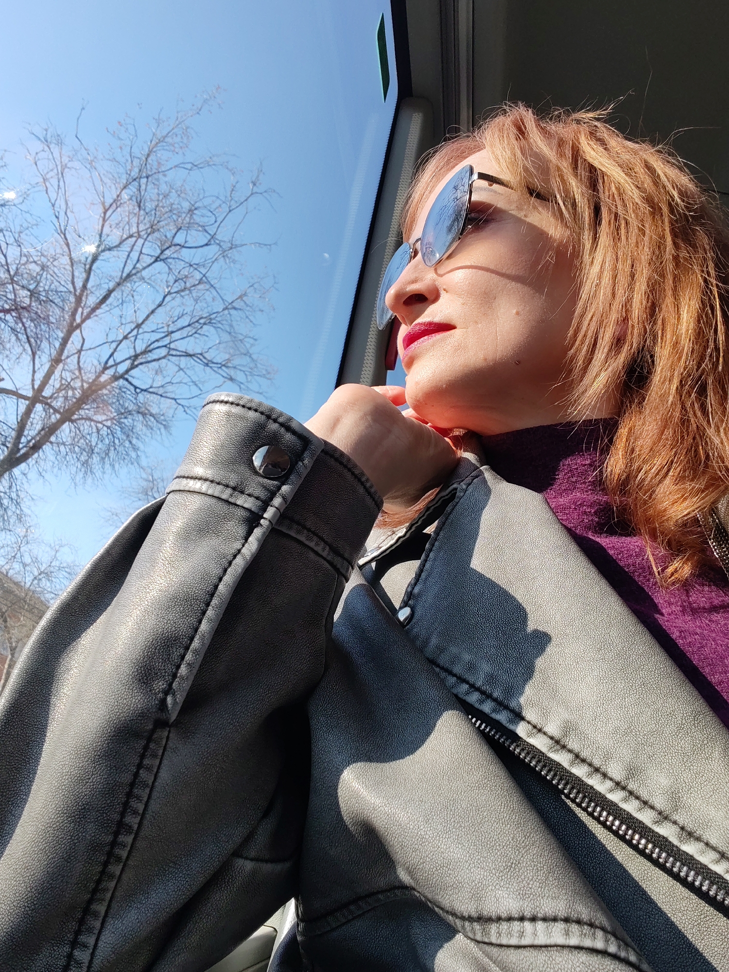 A fair-skinned, middle-aged woman with auburn hair and red lipstick is captured in a medium shot, seated and looking out a window. She is wearing reflective sunglasses, a dark purple turtleneck sweater, and a light gray leather jacket. Her left hand is raised to her chin in a pensive gesture. Outside the window, a clear blue sky and bare tree branches are visible. The photo was taken on a sunny day in the city of Chaikovsky, Russia.
