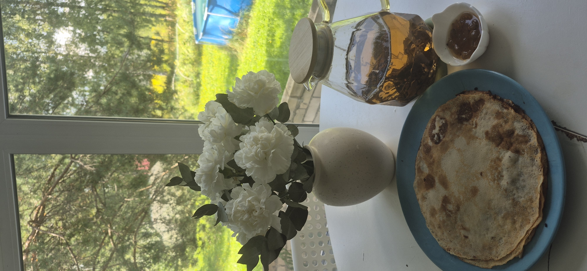 In the foreground of the image, a blue plate holds a stack of crepes. To the right of the plate, a small white dish contains a jam-like substance. Behind the plate and dish, a glass teapot filled with amber-colored tea sits on a white table. The teapot has a wooden lid and a curved handle. To the left of the teapot is a light-colored vase holding a bouquet of white flowers, possibly carnations, with green foliage.
The table is positioned near a window, through which a green, leafy background is visible. The scene appears to be set in a yard with trees and a blue inflatable swimming pool in the distance. The overall impression is that of a breakfast or tea setting on a bright day, potentially in the village of Bezvodnoye, Russia, given the context provided, though this cannot be definitively confirmed from the image alone. There are no people visible in the image.