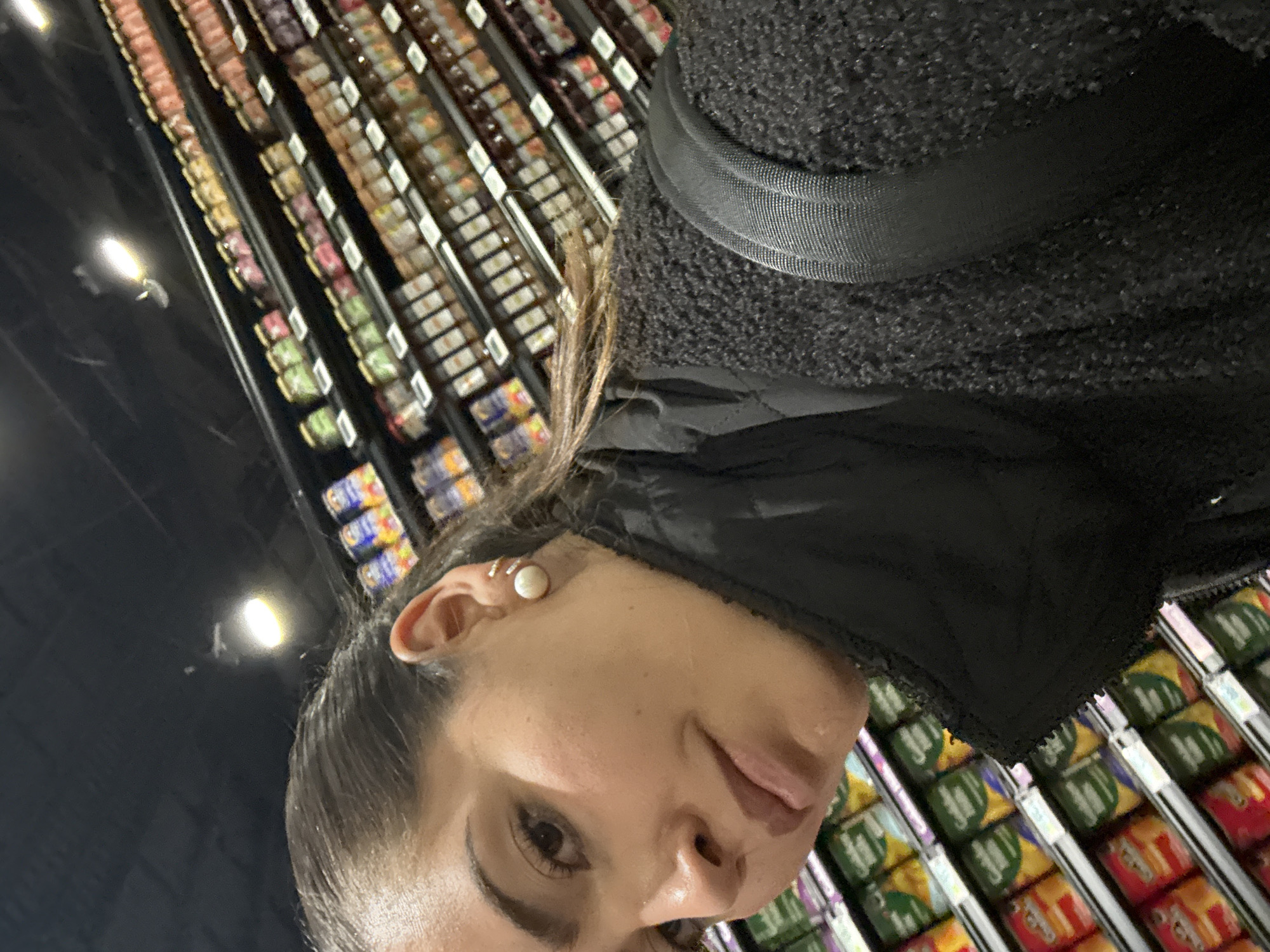 A female individual is centrally depicted in a slightly tilted, close-up portrait within a grocery store aisle, located in Puebla, Mexico. She has dark hair styled away from her face, exposing a large, round, white pearl earring and two small, metallic ear cuffs on her right ear. Her attire consists of a black padded vest worn over a dark, textured, possibly fleece garment. A black strap, consistent with a backpack or shoulder bag, is visible over her right shoulder. Her gaze is directed towards the viewer with a neutral expression. The background features multiple rows of retail shelving laden with various packaged products. To the subject's right, numerous jars with colorful labels and metallic lids, likely containing preserves or sauces, are neatly arranged. To her lower left, shelves display canned goods, including items bearing "Golden" and "Chef Boyardee" branding. The ceiling above is dark, with illuminated recessed lighting fixtures. The scene provides no overt actions or interactions beyond the subject being photographed in a retail environment.