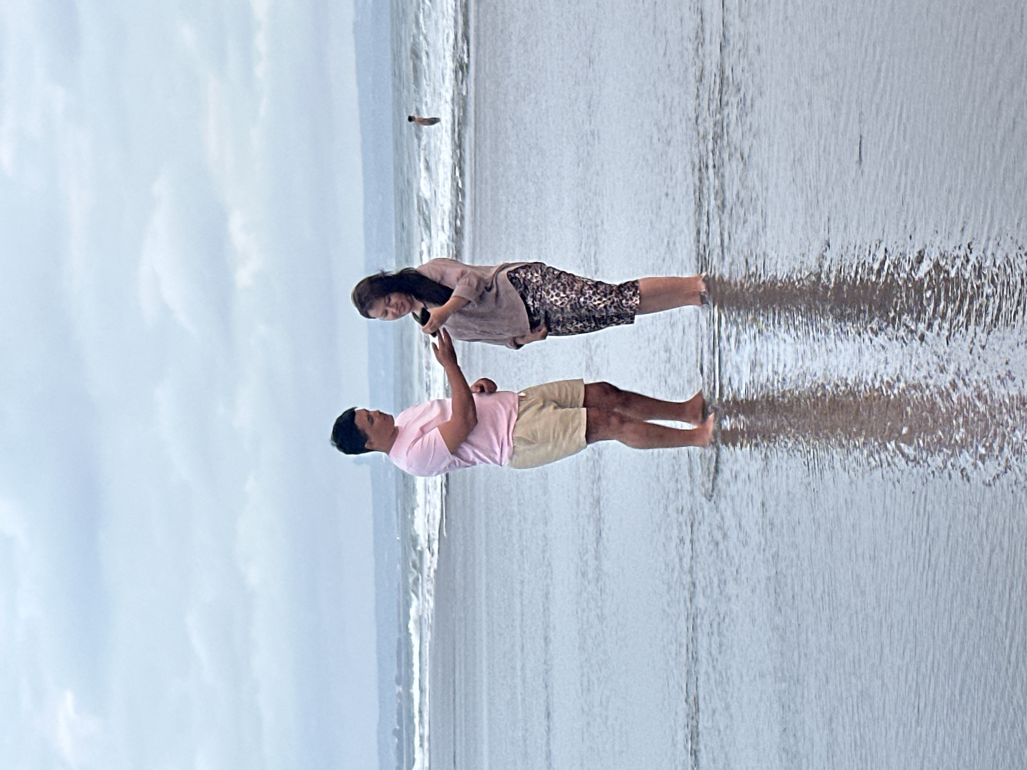 This is a vertical, eye-level shot of two people standing in shallow water at a beach. The man in the foreground is wearing a pink t-shirt and beige shorts, and he is looking down at his phone. The woman behind him is wearing a mauve top and patterned shorts, and she is also looking at her phone. They are standing close together, and their reflections are visible in the water. The background shows a wavy ocean with a cloudy sky. The scene appears to be in North Kuta, Indonesia, and the lighting suggests it might be daytime. The overall mood is relaxed and intimate. There is no visible text in the image.