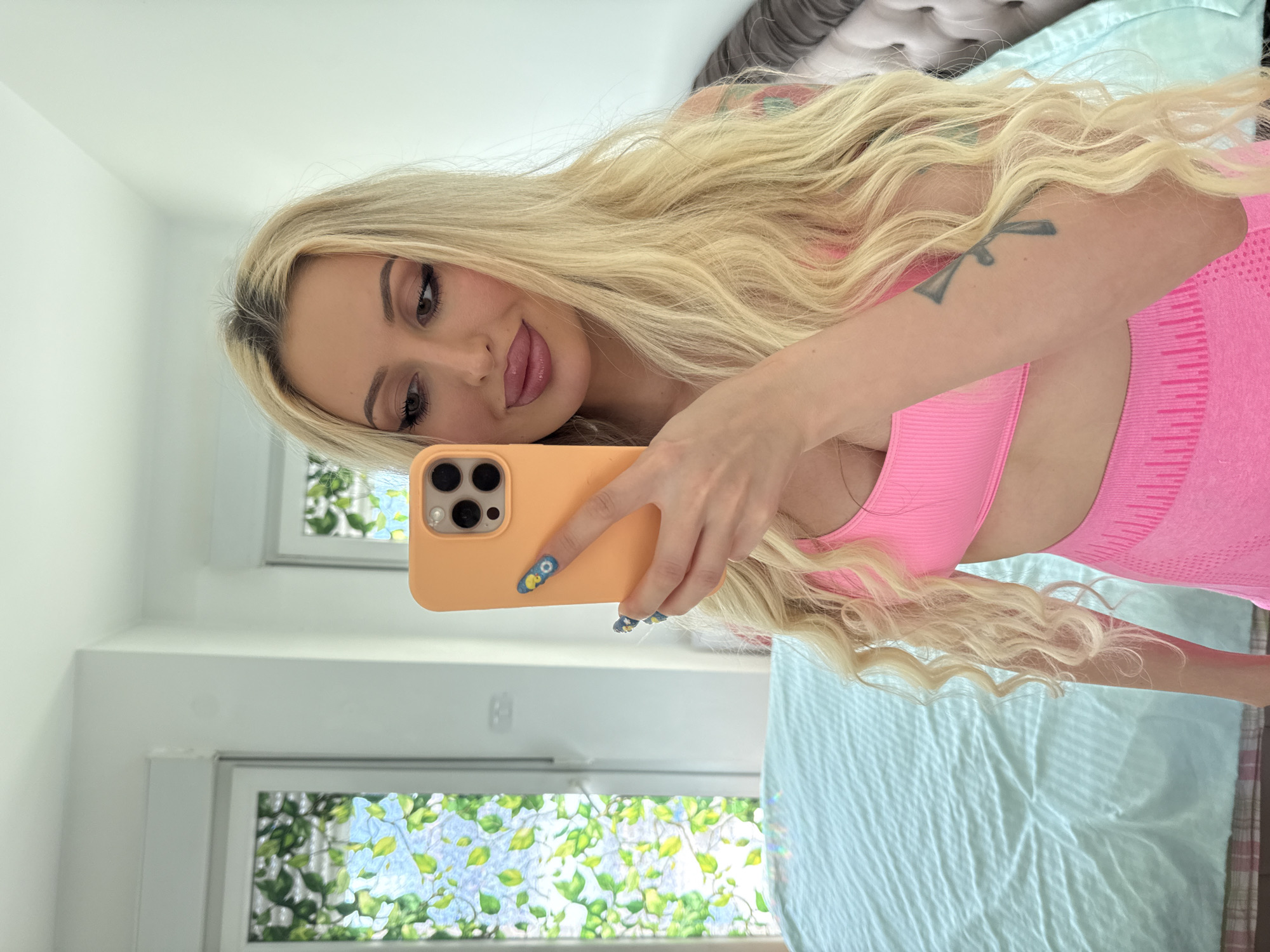 A blonde woman is taking a selfie in a room. She is wearing a pink sports bra and pink shorts. Her hair is long and wavy. She has makeup on and long eyelashes. Her nails are painted with blue and yellow designs. She is holding an orange cell phone in her right hand. In the background, there are two windows with stained glass depicting green leaves. A light blue-green comforter is on a bed to the left. To the right of the woman, a tufted headboard is visible. The lighting in the room is bright, suggesting it is daytime.