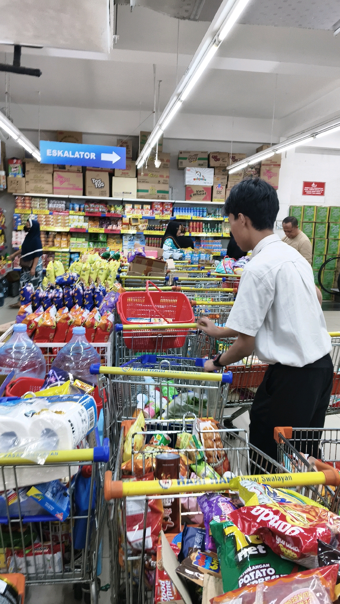 The image shows the interior of a brightly lit supermarket in West Jakarta, Indonesia. A young man in a white short-sleeved shirt and black pants is pushing a shopping cart filled with groceries. In front of him, a red shopping basket is placed in another cart. The shelves in the background are stocked with various food items and household products. Two women wearing hijabs are also visible in the background, one examining products on a shelf and the other near the checkout area. An "ESKALATOR" (escalator) sign with an arrow pointing to the right is displayed overhead. Several shopping carts, some filled with purchases and others empty, are arranged throughout the aisle. The overall scene depicts a typical shopping experience in a modern Indonesian supermarket.