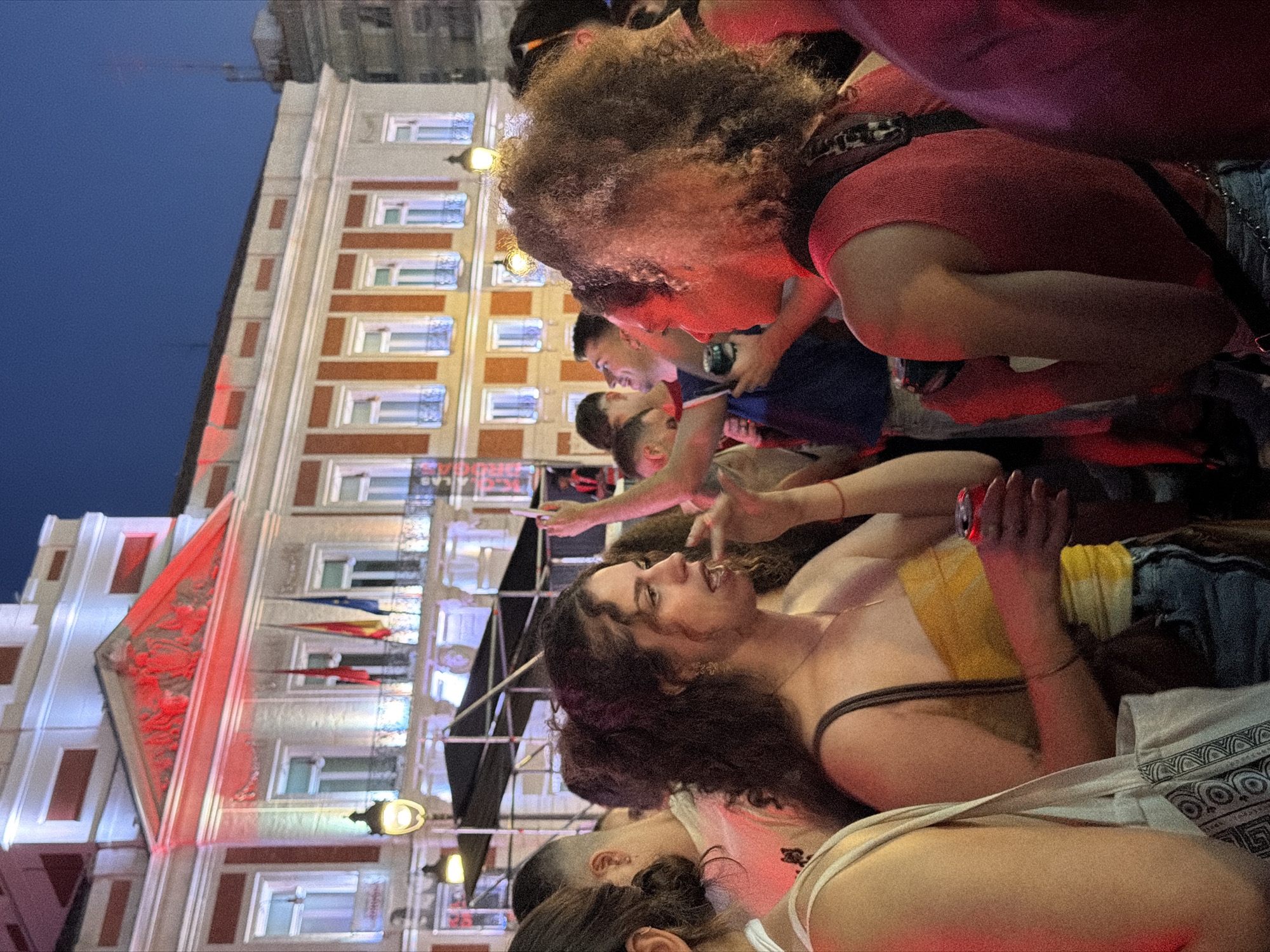 The image depicts a crowd of people gathered in front of a building illuminated with red and white lights. In the foreground, two women are prominent. The woman on the left has curly brown hair and is wearing a yellow and white strapless top and denim shorts, holding a red can in her right hand. The woman on the right has curly hair and is wearing a red tank top. Other individuals are visible in the background, some holding up their phones. The building appears to be a large, ornate structure with multiple windows and flags displayed. The scene suggests an outdoor event or gathering at dusk or night. Based on the architecture of the building, the flags, and the context of large public gatherings, the location is likely the Puerta del Sol in Madrid, Spain.