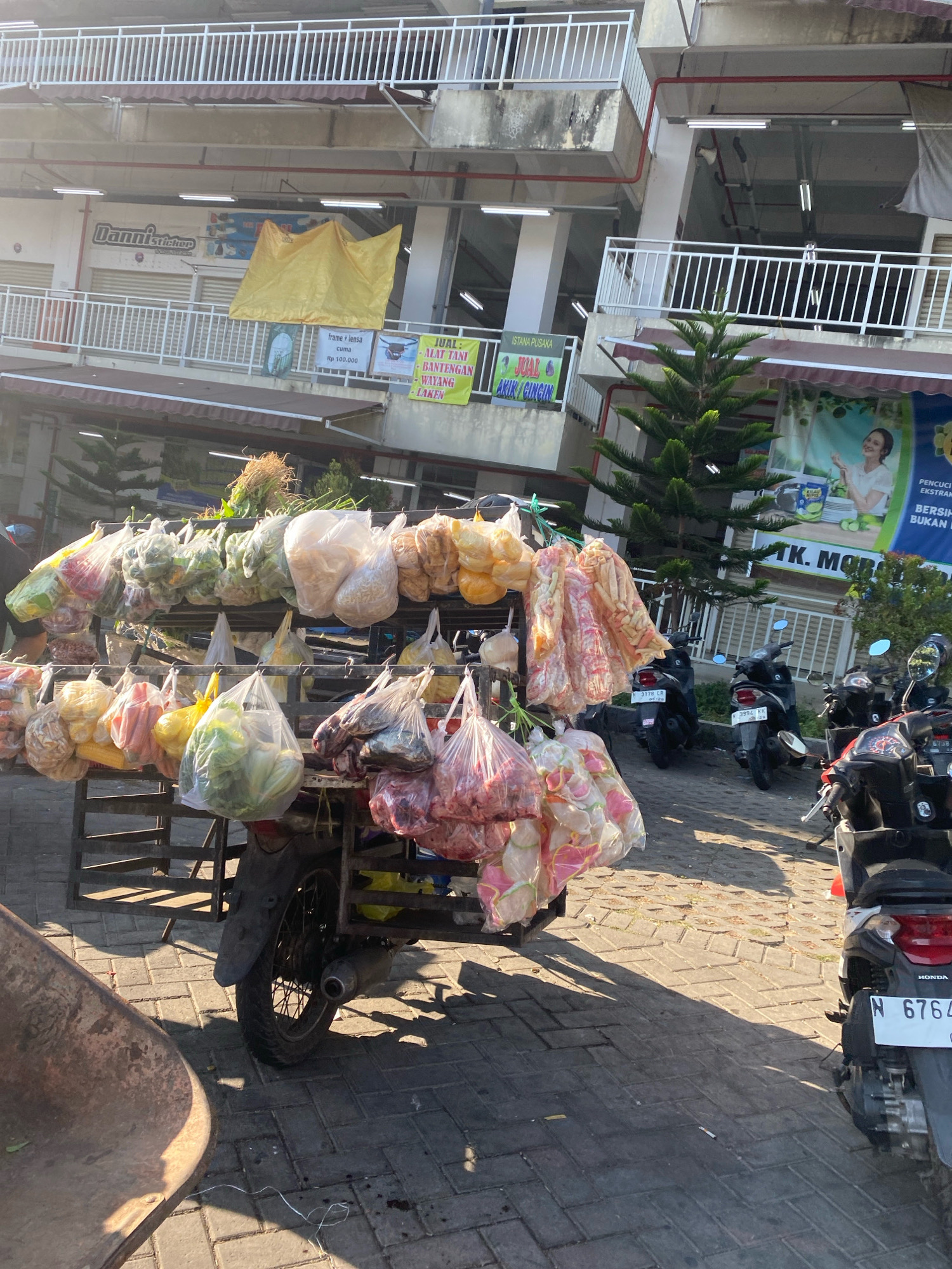 An outdoor marketplace scene is depicted in daylight, with a focus on a vendor's cart laden with various goods. The cart, which appears to be attached to a motorcycle, is overflowing with produce and packaged items. In the foreground, a rusty wheelbarrow is visible. Behind the cart, several motorcycles are parked. The background reveals a multi-story building with balconies and awnings, suggesting an urban setting. Signs are visible on the building, some in Indonesian. The ground is paved with interlocking bricks, and shadows indicate the direction of the sun. The overall impression is of a bustling, informal market.